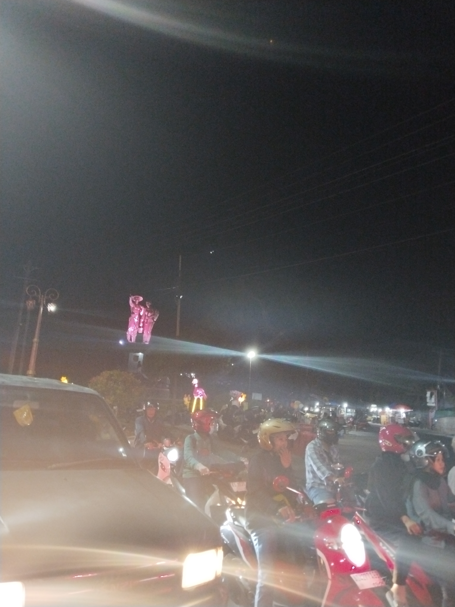 This is a nighttime, low-resolution image of a street scene in Bulakrejo, Indonesia. The foreground is dominated by the hood and headlights of a vehicle, with streaks of light indicating motion or camera shake. In the mid-ground, a group of people on motorcycles are stopped, suggesting a traffic jam or waiting at an intersection. Several individuals are wearing helmets, and some are looking forward. The motorcycles are varied in color and design, with one prominent red scooter. In the background, a large, pink illuminated statue is visible, appearing to be a human figure or group of figures. Streetlights and other distant lights indicate the presence of buildings and infrastructure. The sky is dark, with some faint light beams or lens flares visible. The overall impression is one of a busy urban street at night.