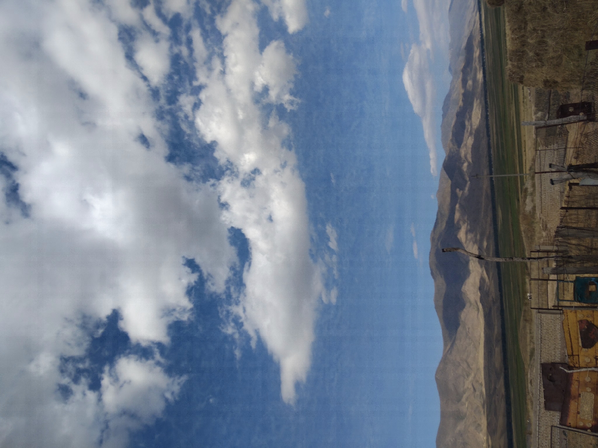 The image depicts a rural landscape under a partly cloudy sky, with mountains in the background. The foreground features a rustic fence made of wire and wooden posts, with some debris including what appears to be rusted metal sheeting and hay bales. A tall, dead tree trunk stands to the right of the fence. Beyond the fence, a vast expanse of green fields stretches towards the undulating, brown and tan mountains, which are partially obscured by shadows from the clouds. The sky is a vibrant blue, dotted with white cumulus clouds, some of which are quite large and billowy. The scene suggests an agricultural setting in a mountainous region, possibly in or around Kopuro Bazar, Kyrgyzstan. There are no people visible in the image.