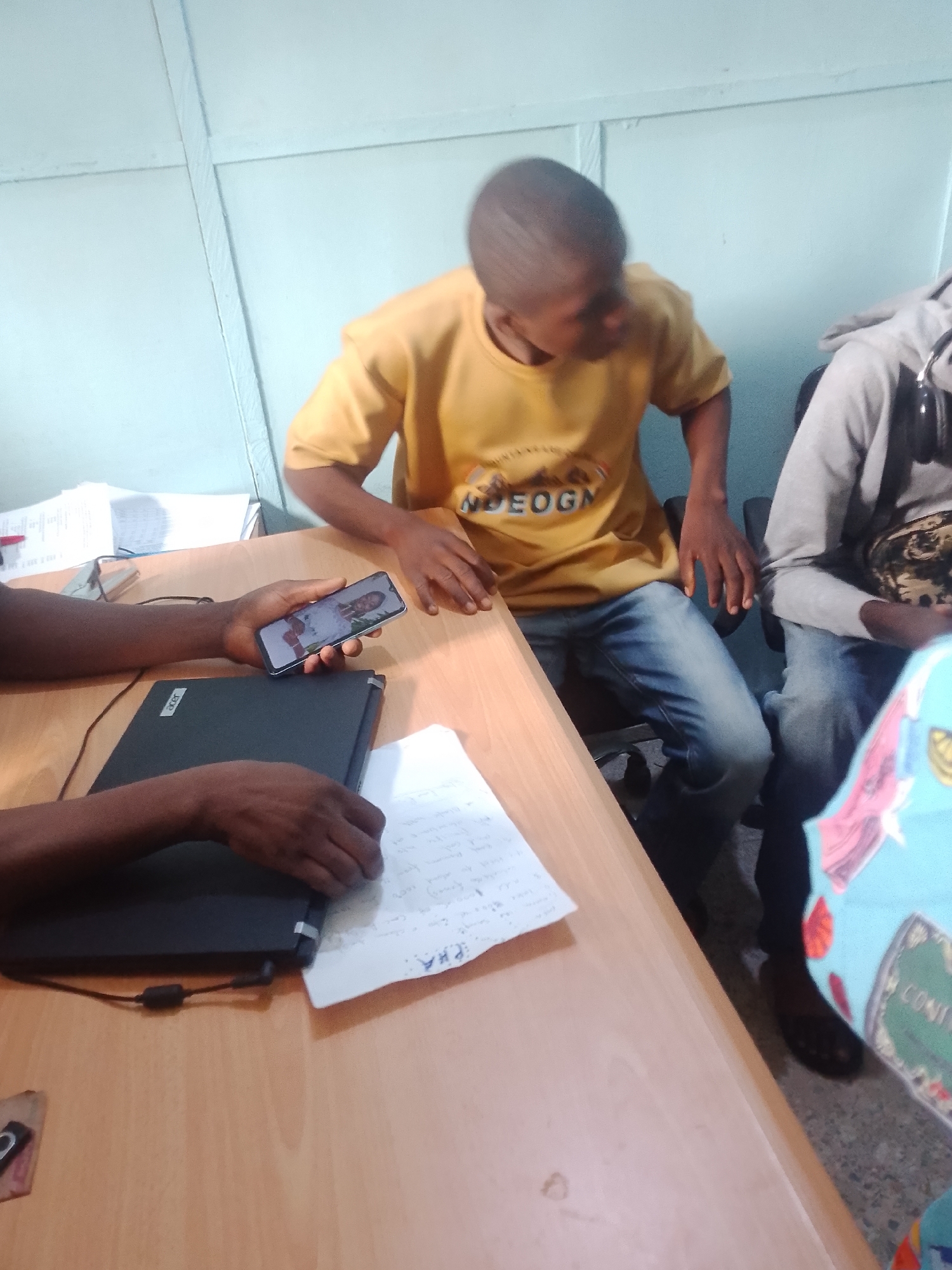 The image shows a group of people in an indoor setting, likely an office or classroom, based on the presence of a desk, laptop, and papers. One person in the foreground is holding a smartphone, displaying a photo of a person. Another person, wearing a yellow t-shirt with "NDEOGN" printed on it, is seated and looking down. To the right, another individual is visible, wearing a grey hoodie and headphones. The scene appears to be casual, with individuals engaged in their own activities. The lighting suggests it could be daytime, but there are no clear external cues like windows. The overall mood seems relaxed and informal. The visible text includes "NDEOGN" on a t-shirt, and some handwritten text on a piece of paper, which is difficult to decipher clearly. There is also text on a printed item on the far right, which reads "CONI".