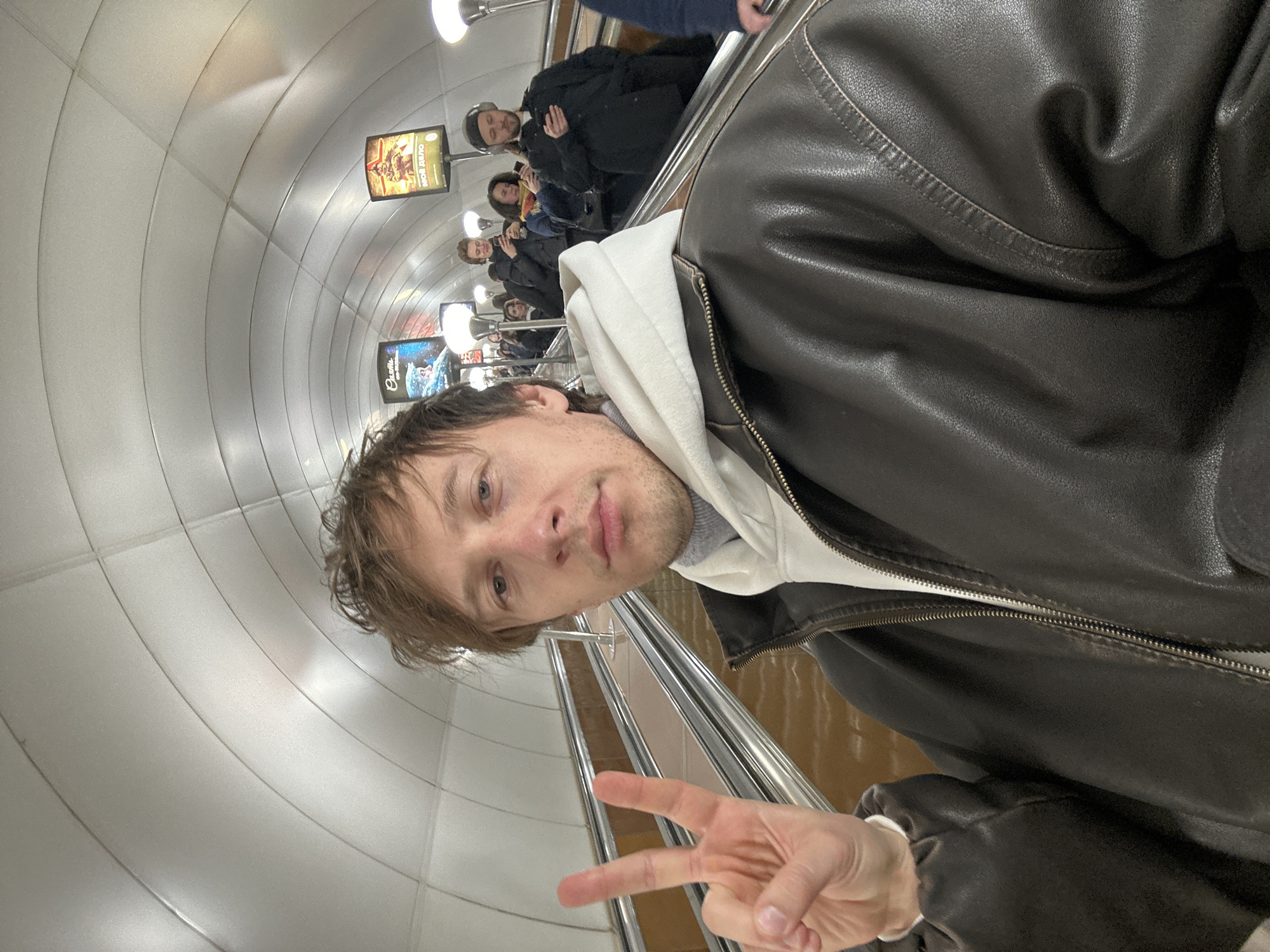 A man in a black leather jacket and white hoodie takes a selfie in a subway station in Saint Petersburg, Russia. The subway has escalators. The man is making a peace sign. A row of people, including at least one wearing a dark-colored hat, is visible in the background on the escalator. There are advertising posters on the wall. The walls of the station are a curved, metallic-colored tile.