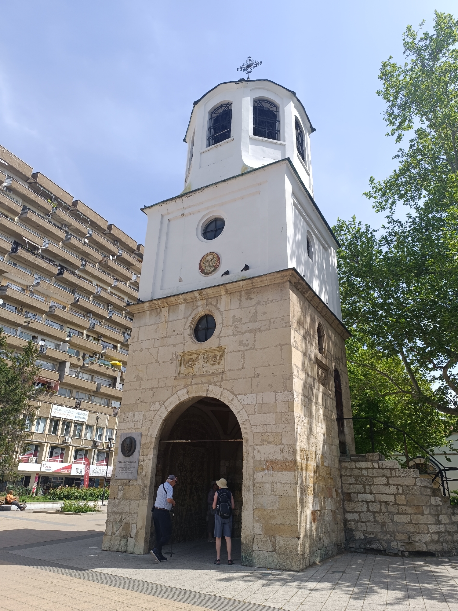 The image shows the Saint George Mausoleum Chapel in Pleven, Bulgaria. The chapel is a tall, multi-level structure made of stone and white-painted sections. The lowest level has an arched entrance, through which people are entering. To the right of the entrance is a set of stone stairs. The top level has arched windows and a cross on top. To the left of the chapel is a tall building with multiple balconies and windows. To the right is a green tree. The ground is paved. Several people are visible, including two entering the chapel and one standing outside of it. A plaque is on the wall near the entrance.