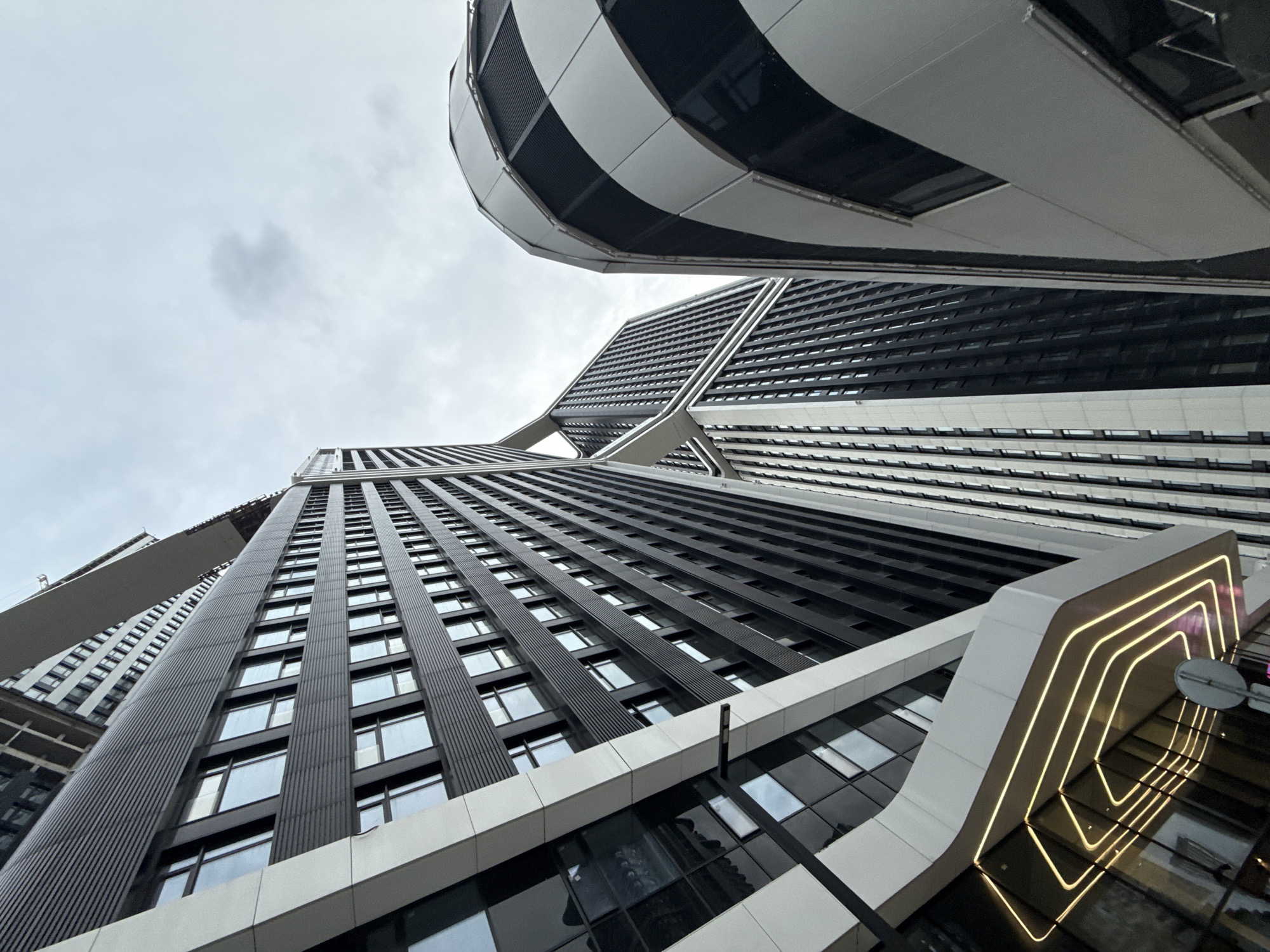 The image is a worm's-eye view of two modern skyscrapers in Moscow, Russia. The buildings feature a pattern of black and white horizontal stripes with windows, and one of the buildings has an elevated, rounded section on top with the same stripe pattern. A section of the other building has a decorative design of recessed, glowing lines. The sky is overcast.