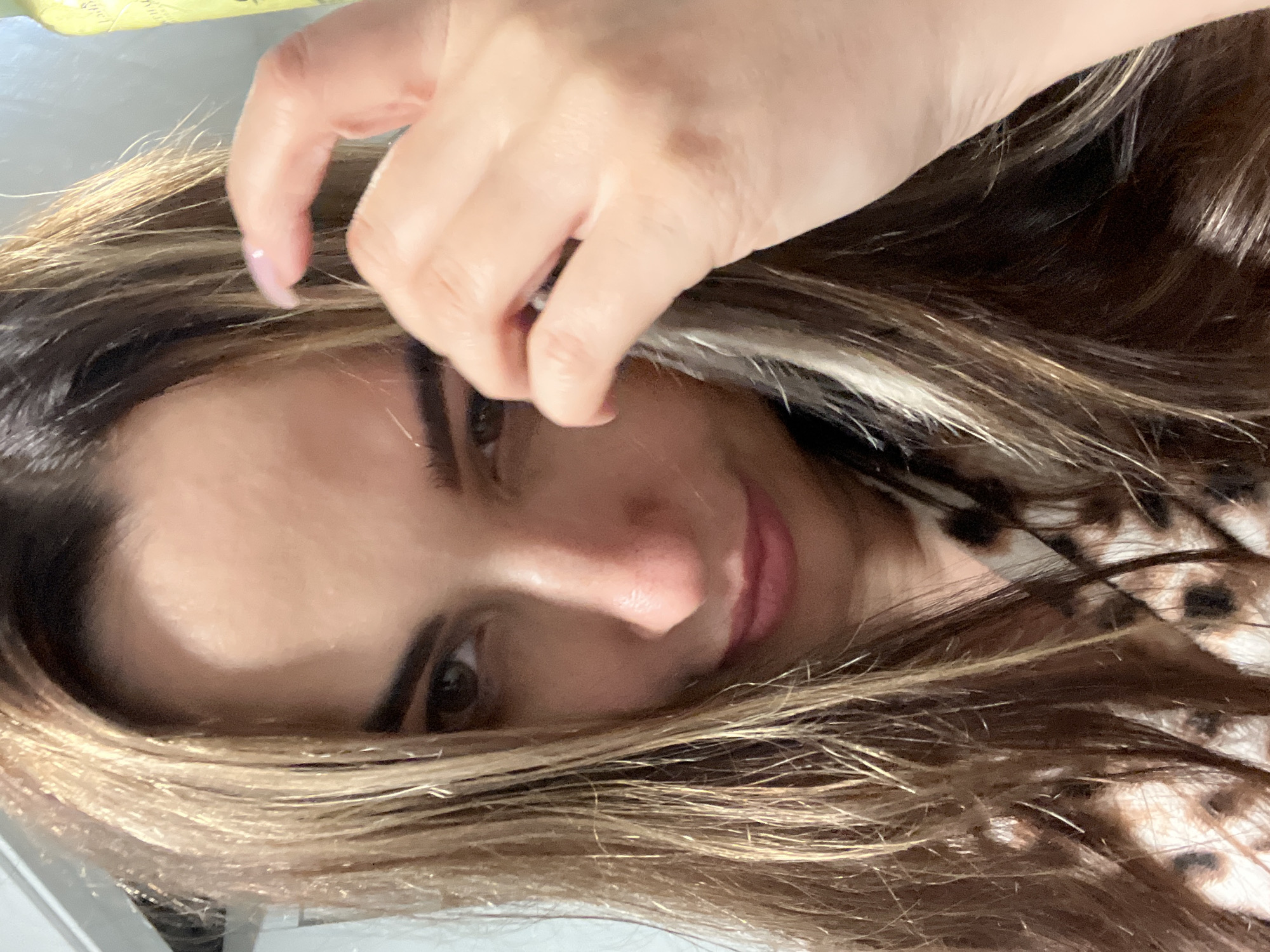 A close-up, head-and-shoulders portrait captures an adult female in an unknown indoor location. The subject has long, wavy, multi-toned hair, predominantly dark brown with significant blonde or light brown highlights, which cascades around her face. Her gaze is directed downward and slightly to her left (viewer's right). Her left eye (viewer's right) is fully visible and dark brown, while her right eye is partially obscured by hair and angle. Her eyebrows are dark and well-defined. The tip of her nose shows a slight reddish tint, and her lips are parted in a subtle, almost imperceptible smile, exhibiting a light pink color. Her right hand (viewer's left) is raised, with fingers gently touching strands of hair near her right temple. The nail on her right index finger is long and appears to have a clear or pale pink coating. She is wearing a garment with a brown, tan, and black animal print pattern, visible around her neck and chest. The background is indistinct; a light-colored, possibly reflective surface is on the left, and the top portion of a tall, narrow, light-green or yellow-green bottle is partially visible on the right. The lighting is soft and even.