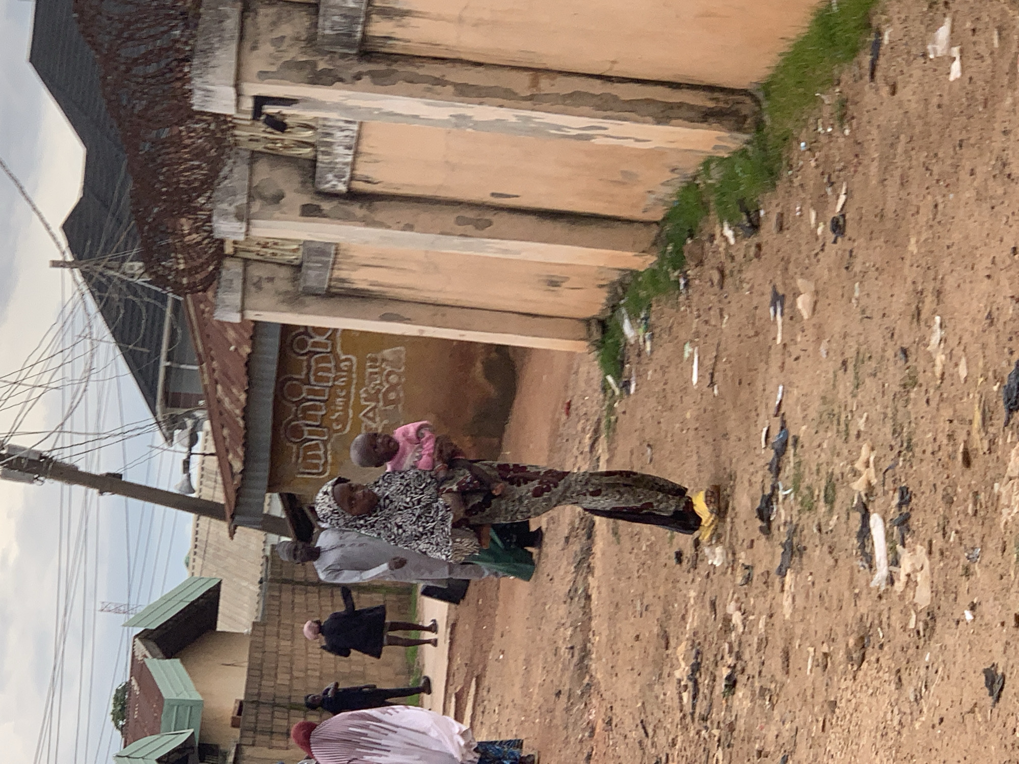 In Jos, Nigeria, a young woman in a black and white headscarf and a patterned dress carries a baby wearing a pink garment. She is walking on a dirt road littered with debris, with a wall adorned with barbed wire to her right. Behind her, a man in a grey kaftan walks, followed by other individuals. The background reveals buildings with corrugated iron roofs and what appears to be a utility pole with numerous wires. The overall scene depicts a street in a moderately populated urban area.