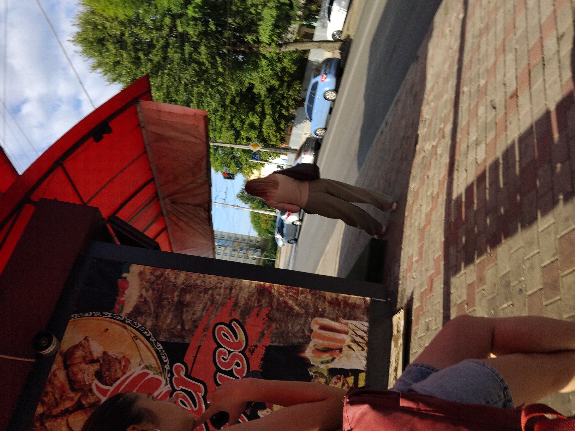 The image shows a street scene in Zhytomyr, Ukraine, on a sunny day. In the foreground, a person wearing a red backpack and denim shorts is standing on a brick sidewalk. To the left, a large advertisement for a food establishment, likely a kebab shop, is visible, featuring images of cooked meat and a shawarma cone. Behind this, a red awning extends over the sidewalk.
Further down the street, a woman with long, reddish-brown hair is walking away from the camera, carrying a dark bag over her shoulder. She is wearing a light brown shirt and loose-fitting olive-green pants. The street is lined with trees, and several cars are parked or driving. Visible vehicles include a light blue station wagon, a silver car, and a white car. In the background, a multi-story building and a traffic light can be seen. The scene suggests a typical urban environment with commercial activity and pedestrian traffic.