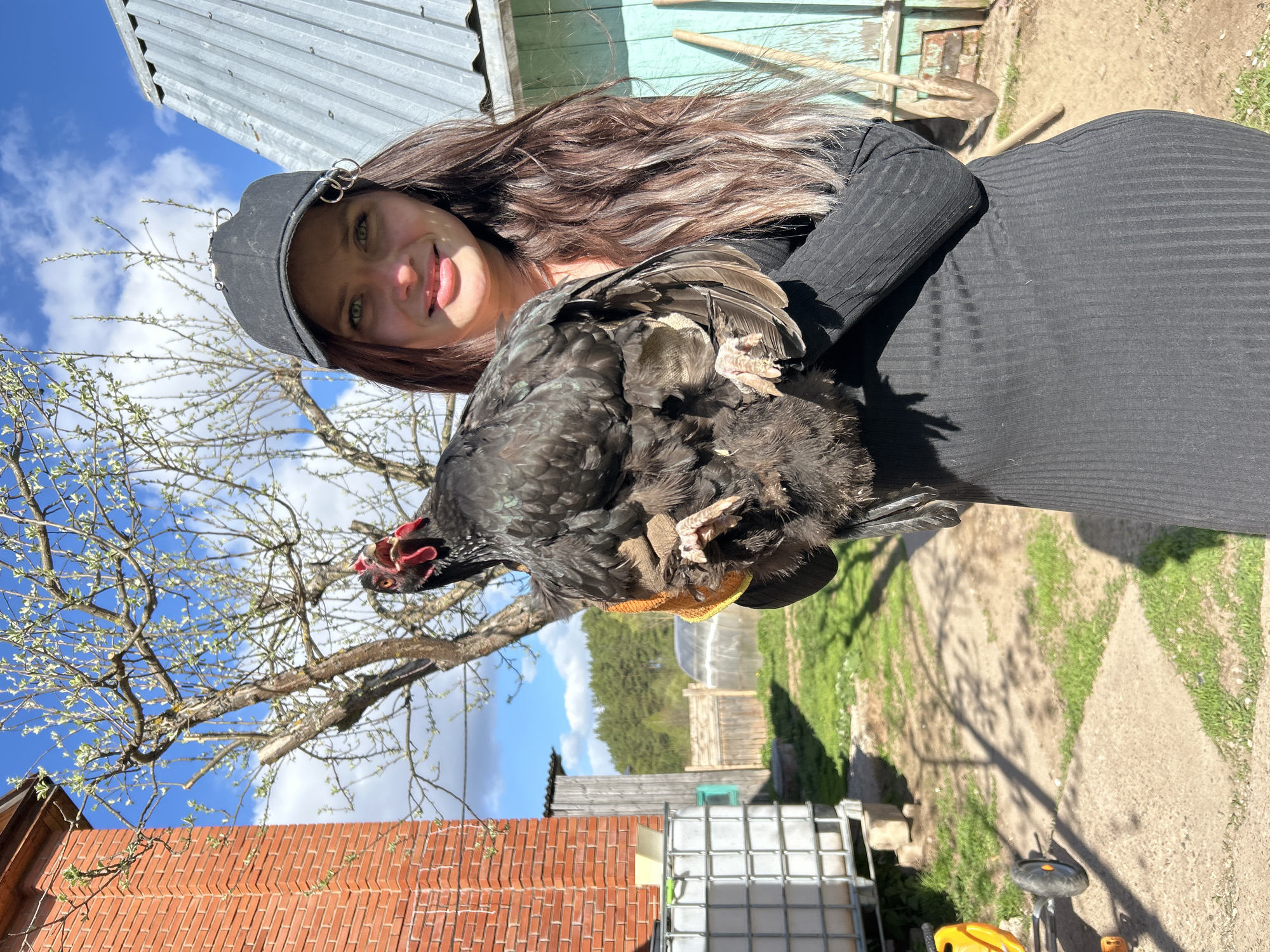 A woman with long, wavy brown hair and green eyes is holding a black chicken. She is wearing a black cap adorned with silver rings and a black long-sleeved dress. The location appears to be in a rural setting, possibly in Rusalevka, Russia. The background includes a brick building, trees with budding leaves, a metal shed, and a blue sky with scattered clouds. There is also a large white container, and some green grass is seen in the background. The woman is smiling.