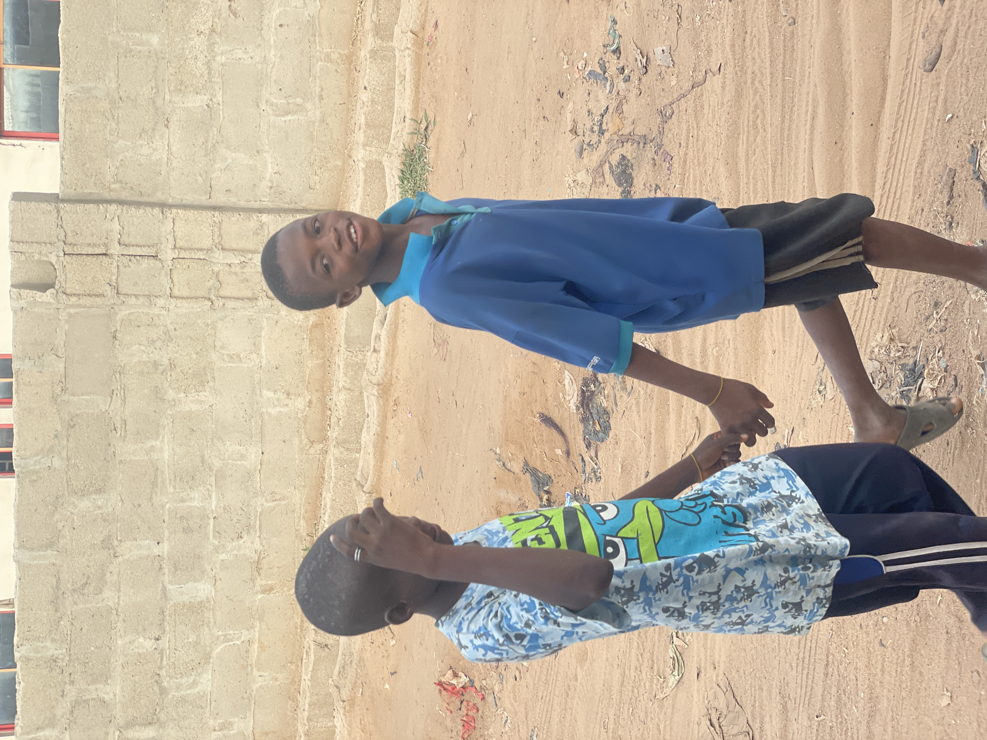 Two young Black males stand holding hands in Lokoja, Nigeria. One wears a blue and light-blue patterned shirt with a cartoon image, dark blue pants with white stripes, and green sandals. The other wears a blue shirt, black shorts with white stripes, and is barefoot. They stand on sandy ground littered with debris. A tan brick wall with red window frames is visible on the left.