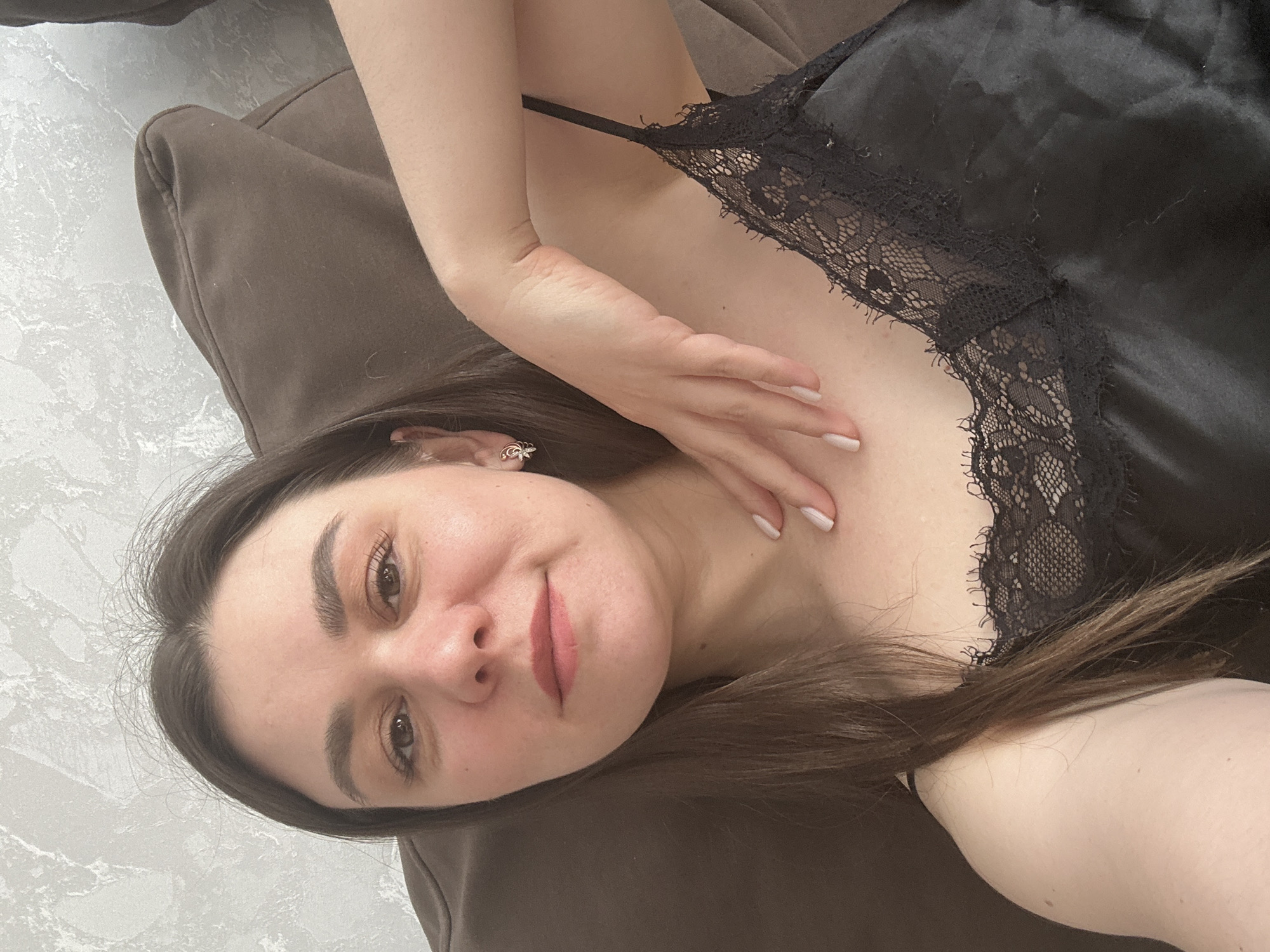 A portrait image, rotated 90 degrees counter-clockwise, depicts a young woman in an indoor setting. She is lying down, with her head resting on a soft, brown surface to the left of the frame, and her body partially visible extending towards the right. She appears to be in her 20s or early 30s, with long, dark brown hair that cascades around her shoulders. Her face, turned towards the camera, shows a gentle, closed-mouth smile. She has dark eyebrows, brown eyes, and is wearing light makeup, including soft pink lipstick. A small, decorative earring is visible in her right ear.
She is dressed in a black satin top or camisole, adorned with intricate black lace along the neckline and straps. Her right hand, adorned with light-colored nail polish, is positioned delicately near her chest. She seems to be resting comfortably on what appears to be a brown sofa or cushion. In the background, to the upper left, a light-toned wall with a subtle, textured pattern, possibly simulating marble or stone, is visible. The image has the candid feel of a selfie or a casual photo. This scene is set in Chaikovsky, Russia. No text is visible in the image.