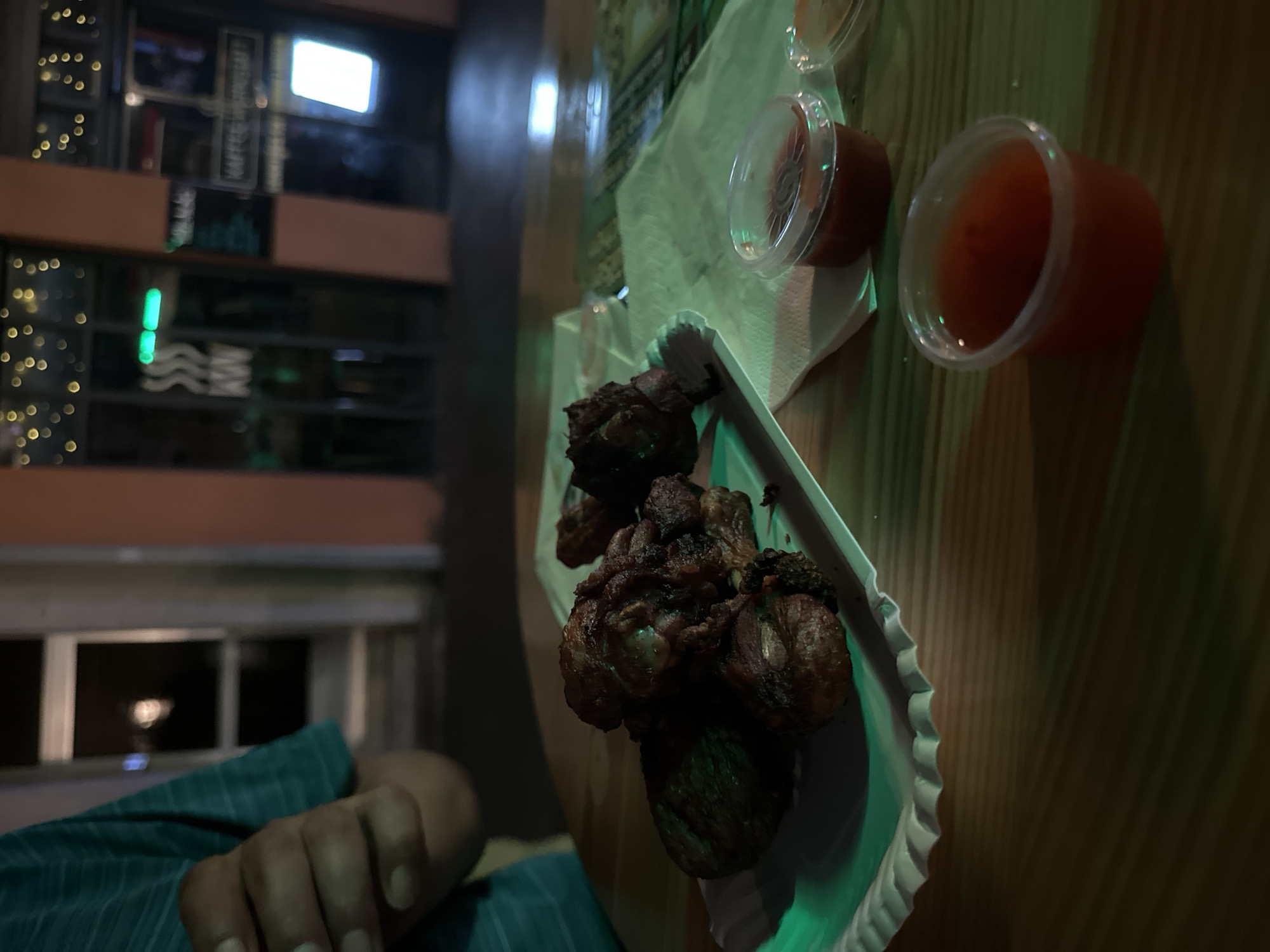 This image captures a close-up view of fried chicken wings served on a white paper plate, placed on a wooden table. Two small clear plastic containers filled with dipping sauce are also visible on the table, one closer to the viewer and another further away, partially obscured by a napkin. In the background, a blurry outdoor setting is suggested, with lights and building facades, potentially indicating a restaurant or bar area. The overall lighting suggests an evening or nighttime scene. There is no specific indication that this location is in Krefeld, Germany, nor any explicit details about people or interactions beyond a person's arm and hand visible on the left side of the frame.
