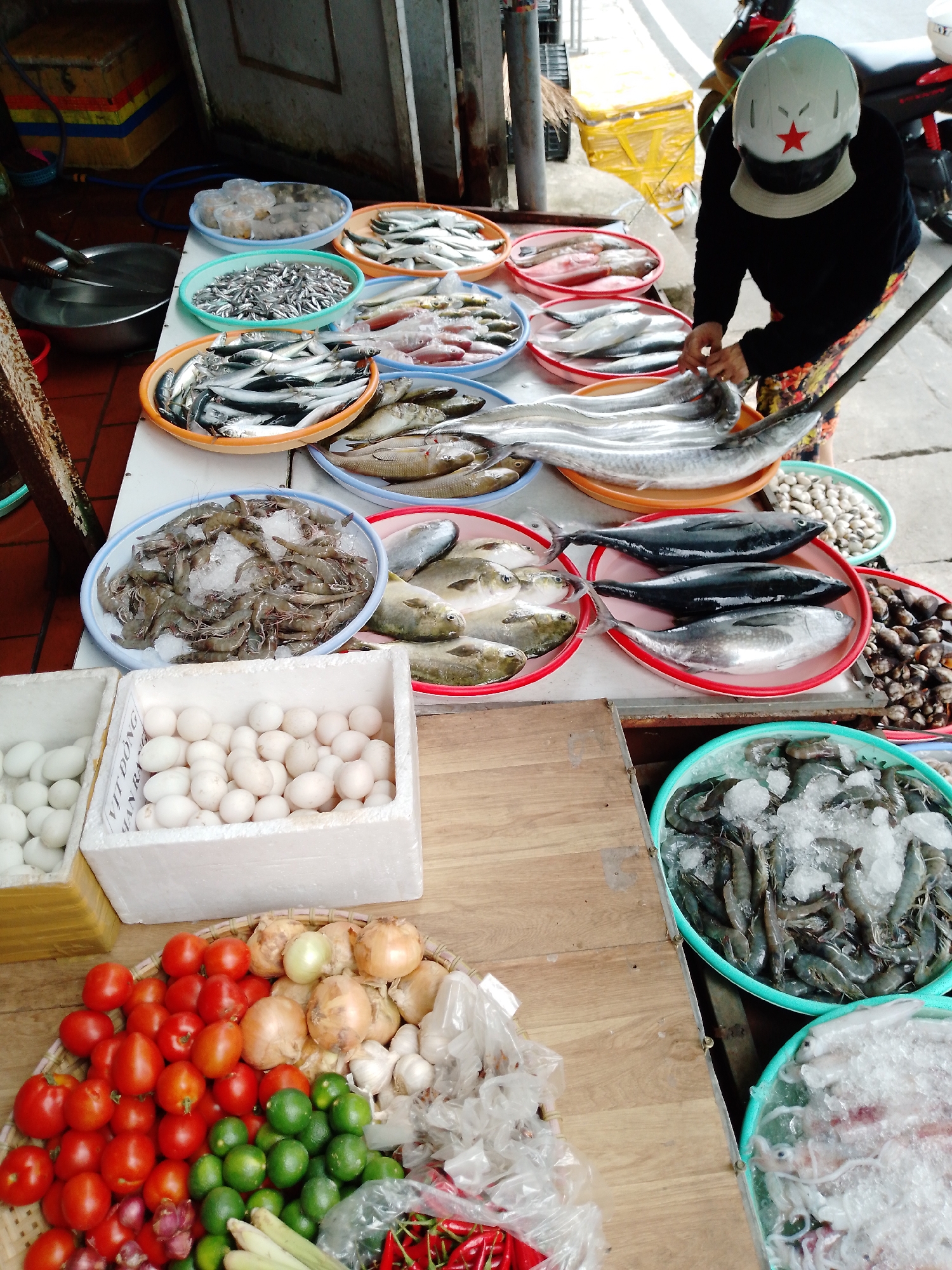 This image captures a vibrant outdoor market stall, likely in Da Lat, Vietnam. The main subject is a vendor, partially visible from the back, wearing a black long-sleeved shirt, patterned pants, and a white helmet adorned with a red star. The vendor is actively engaged with the products, seemingly arranging or preparing the seafood displayed on a long white table.
The stall is laden with a wide variety of fresh goods. The upper part of the table is dominated by an extensive selection of seafood. Numerous shallow, colorful plastic dishes and bowls hold different types of fish, including larger whole fish like kingfish or barracuda, various smaller schooling fish, and flatfish, many resting on ice. Other bowls contain fresh shrimp on ice, clams, and what appears to be squid also on ice.
Below the seafood display, several crates are filled with white eggs, specifically duck eggs as indicated by the partially visible text "VỊT ĐỒNG" on one of the white foam cartons. Adjacent to these, a large woven tray holds an assortment of fresh produce, including bright red tomatoes, yellow onions, green limes, heads of garlic, and red chili peppers, with stalks of lemongrass also visible.
The setting suggests a bustling daytime market, with bright natural lighting illuminating the fresh produce. In the background, part of a building and a red motor scooter can be seen, typical of an urban or semi-urban market environment. The scene depicts a daily commercial activity centered around fresh food sales.