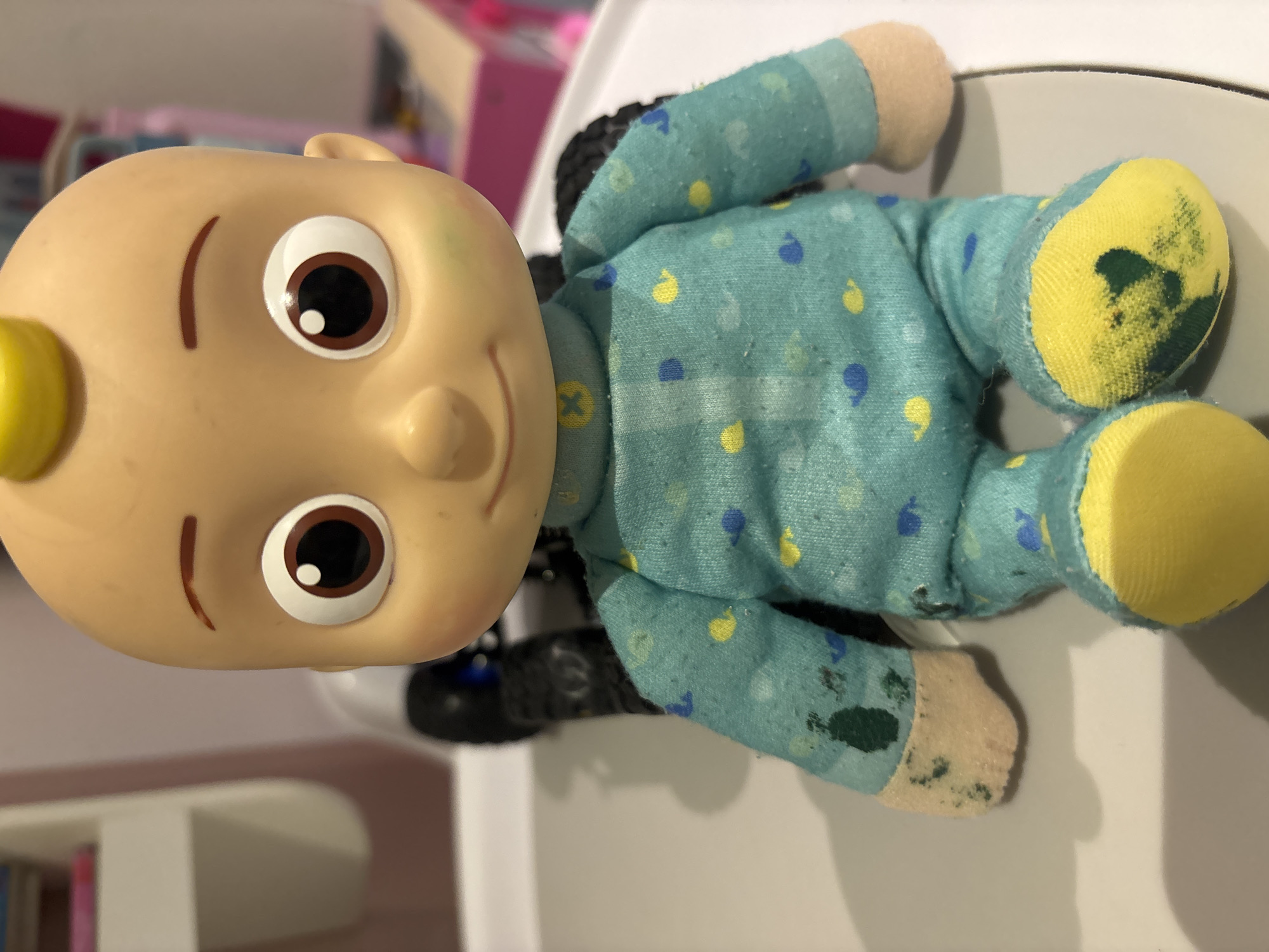 A close-up, eye-level, indoor shot shows a Cocomelon JJ doll sitting on a light grey surface, possibly a tray or table. The doll is facing forward, with its head slightly tilted. Its pale skin has some faint green marks on the left cheek. The doll's large brown eyes, with white pupils, are wide open. It has a small, closed mouth, and a thin brown eyebrow arched over its left eye. A small yellow tuft of hair is visible on its bald head.
The doll is wearing a light blue pajama set adorned with yellow and blue whale and cloud-like patterns. The sleeves of the pajama top show some dark green smudges, particularly on the right sleeve. The doll's fabric hands are a light tan color, and both are smudged with dark green and other dark marks. The doll's legs are also covered by the pajama bottoms, which have the same pattern. The doll's yellow feet are heavily marked with dark green smudges. Behind the doll, a black object with a ribbed texture is visible, possibly a stroller or a toy. In the blurred background, there is a white shelf unit to the left and a pink object, possibly a toy or a piece of furniture, to the right. The lighting is soft, casting a shadow of the doll onto the surface. The image does not provide any information about the city of Dubai, United Arab Emirates.