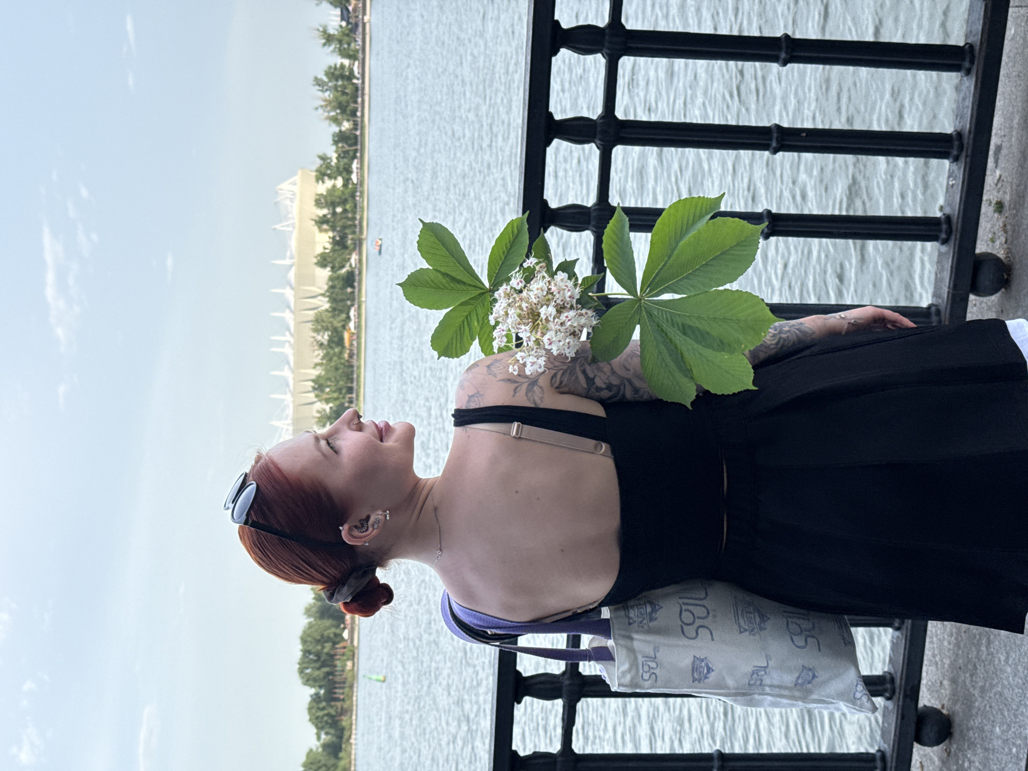 A photograph depicts a woman holding chestnut flowers while standing by a fence. She faces left, toward a river with a cityscape visible in the background. The woman is dressed in a black top and skirt. She has red hair pulled back in a bun and sunglasses pushed back on her head. Tattoos are visible on her arm and shoulder. She is wearing a necklace and multiple earrings. A tote bag hangs from her shoulder. The fence is black wrought iron. The river's surface is rippled. The cityscape includes green trees and tall buildings. This is most likely the city of Rostov-on-Don, Russia.