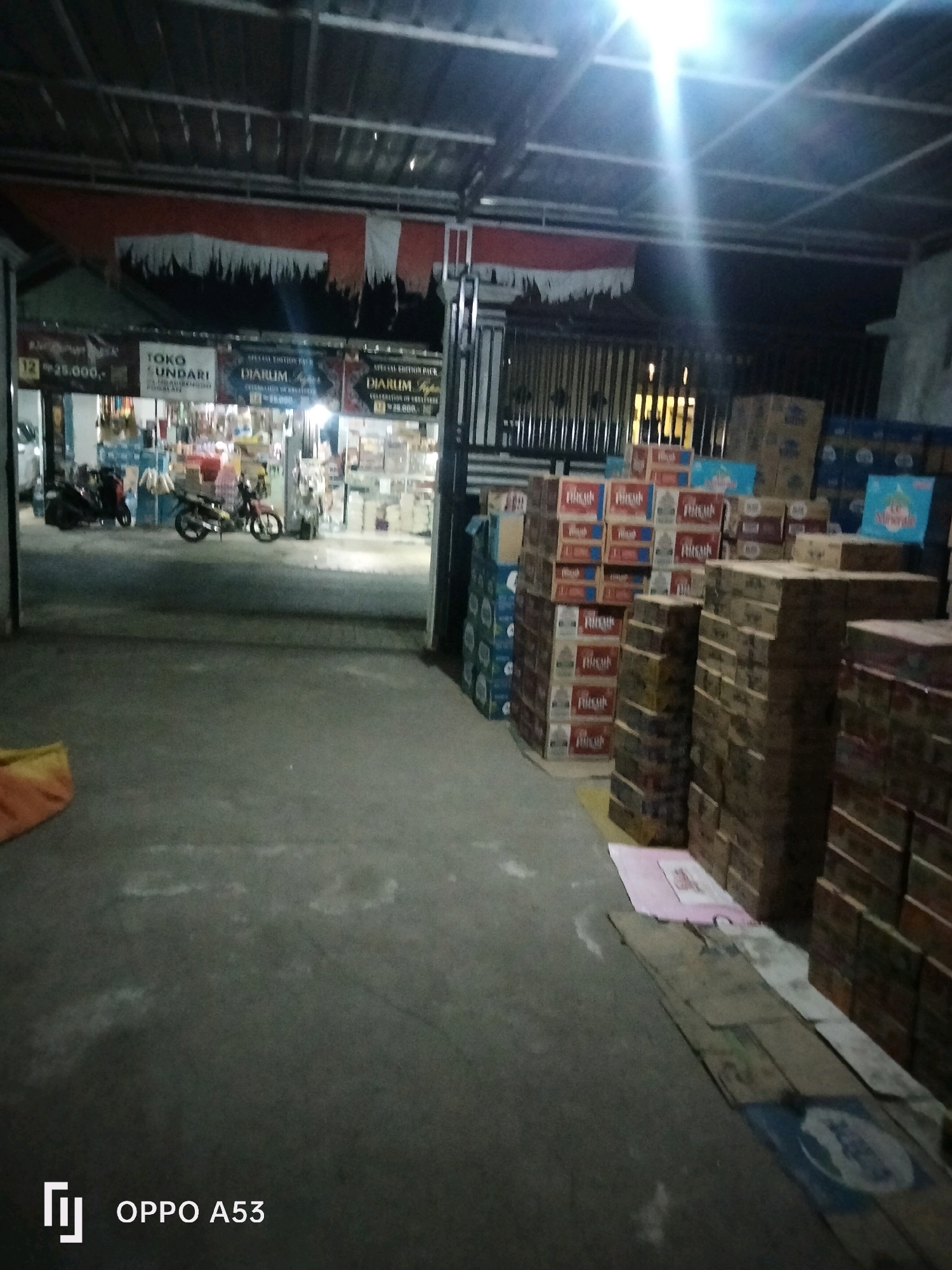 The image shows a nighttime scene in what appears to be a store or warehouse in Trenggalek, Indonesia. In the background, a shop with the signage "TOKO UNDARI" is visible, displaying various goods on shelves. Two motorcycles are parked outside the shop. The foreground is a concrete floor with stacks of boxes to the right, some of which appear to be beverages. The overall lighting suggests artificial illumination from overhead. The environment seems to be an outdoor or semi-outdoor area, possibly an alleyway or a loading dock adjacent to a commercial establishment.