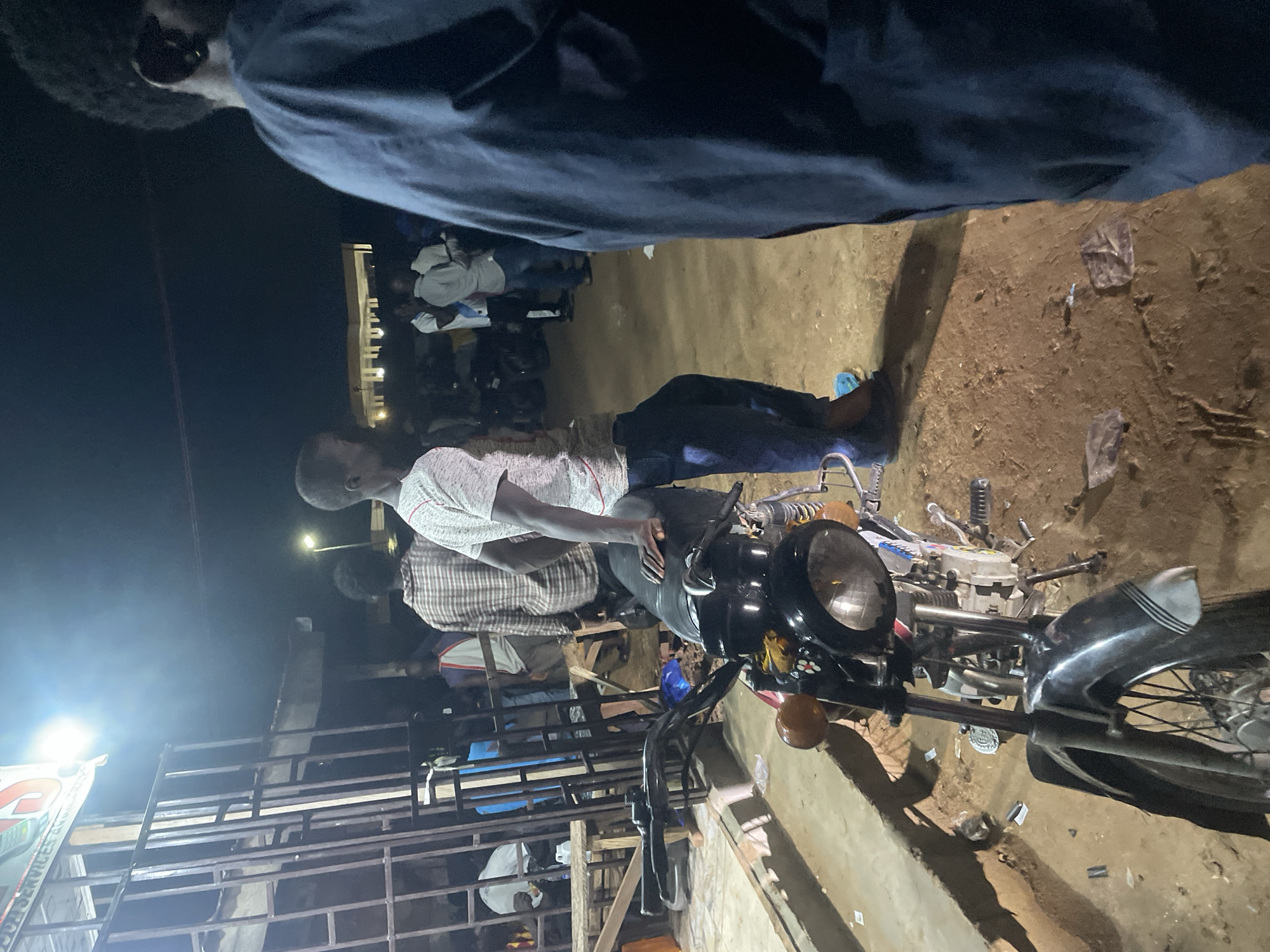 This nighttime photograph, taken in Lokoja, Nigeria, depicts a street scene with several individuals gathered around a parked motorcycle. The foreground is dominated by the front of a black motorcycle, its headlight illuminated. To the left, a metal barrier, possibly part of a shop or stall, is visible.
In the middle ground, a young man in a light-colored, patterned t-shirt and dark pants leans against the motorcycle, looking towards the right. Behind him, other individuals can be seen, some standing and interacting, others in the background. One person wears a plaid shirt, and another a white t-shirt. The environment appears to be an outdoor, possibly dusty or unpaved area, with some scattered debris on the ground. The lighting suggests it's evening or night, with artificial light sources illuminating the scene. A sign with text, partially obscured by glare, is visible in the upper left. The overall atmosphere suggests a casual gathering or public space at night.