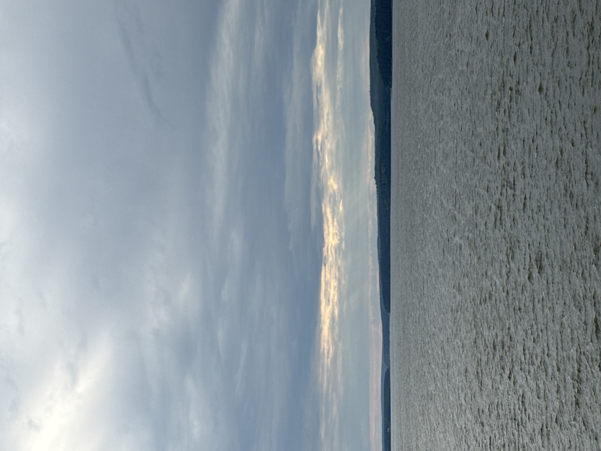 A wide, calm body of water stretches across the lower half of the frame, its surface rippled by a gentle breeze. The water appears to be a lake or river, reflecting the overcast sky. In the distance, a dark, tree-covered shoreline forms a continuous, undulating line against the horizon. The sky above is a canvas of gray clouds, with a bright, sunlit band breaking through in the middle, casting ethereal rays downwards. The overall atmosphere is serene and slightly somber, suggesting a cool or temperate climate. This scene depicts the Votkinskoye Reservoir in Votkinsk, Russia, a prominent landmark that serves as a source of water and a popular recreational area.