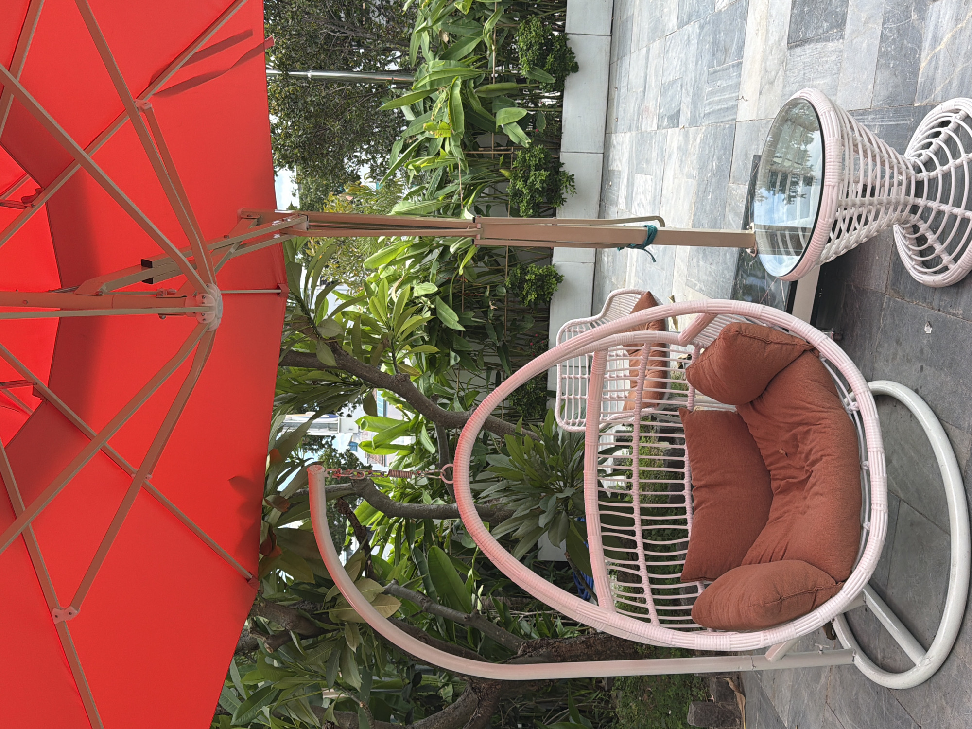 This image depicts an outdoor seating area, likely a patio or balcony, in the city of Đà Nẵng, Vietnam. A large, bright red umbrella provides shade over the scene. Beneath the umbrella, a white wicker hanging chair with rust-colored cushions is prominently featured on the left. To the right of the hanging chair, a small, matching white wicker side table with a glass top is visible. The ground is paved with gray stone tiles. In the background, lush green foliage and plants are arranged in white planters, suggesting a tropical or subtropical environment. The overall impression is one of a relaxing outdoor space, possibly at a resort, hotel, or private residence. There are no people visible in the image.