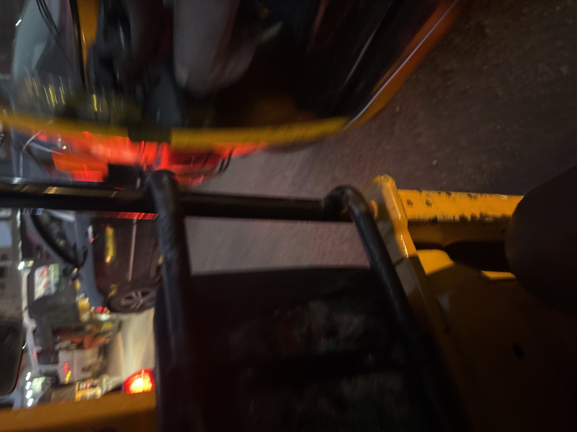 The image depicts a congested street scene in Onitsha, Nigeria, likely during traffic or at night, given the illuminated vehicle lights and overall dim lighting. The perspective is from inside a vehicle, possibly an auto-rickshaw, looking outwards. In the foreground, the bright yellow metalwork of the vehicle is visible, along with a dark, padded seat.
The middle ground is dominated by other vehicles in traffic. A dark-colored car, likely a sedan, is prominently featured, with its taillights glowing red. To its left, an auto-rickshaw with a canvas enclosure, possibly carrying passengers, is also visible. Further back, a white van or bus is partially seen, and other vehicles create a sense of dense traffic. The road surface appears to be asphalt.
The background shows buildings with some illuminated windows, suggesting an urban environment. The overall impression is one of a busy, bustling city street experiencing typical traffic conditions in Onitsha.