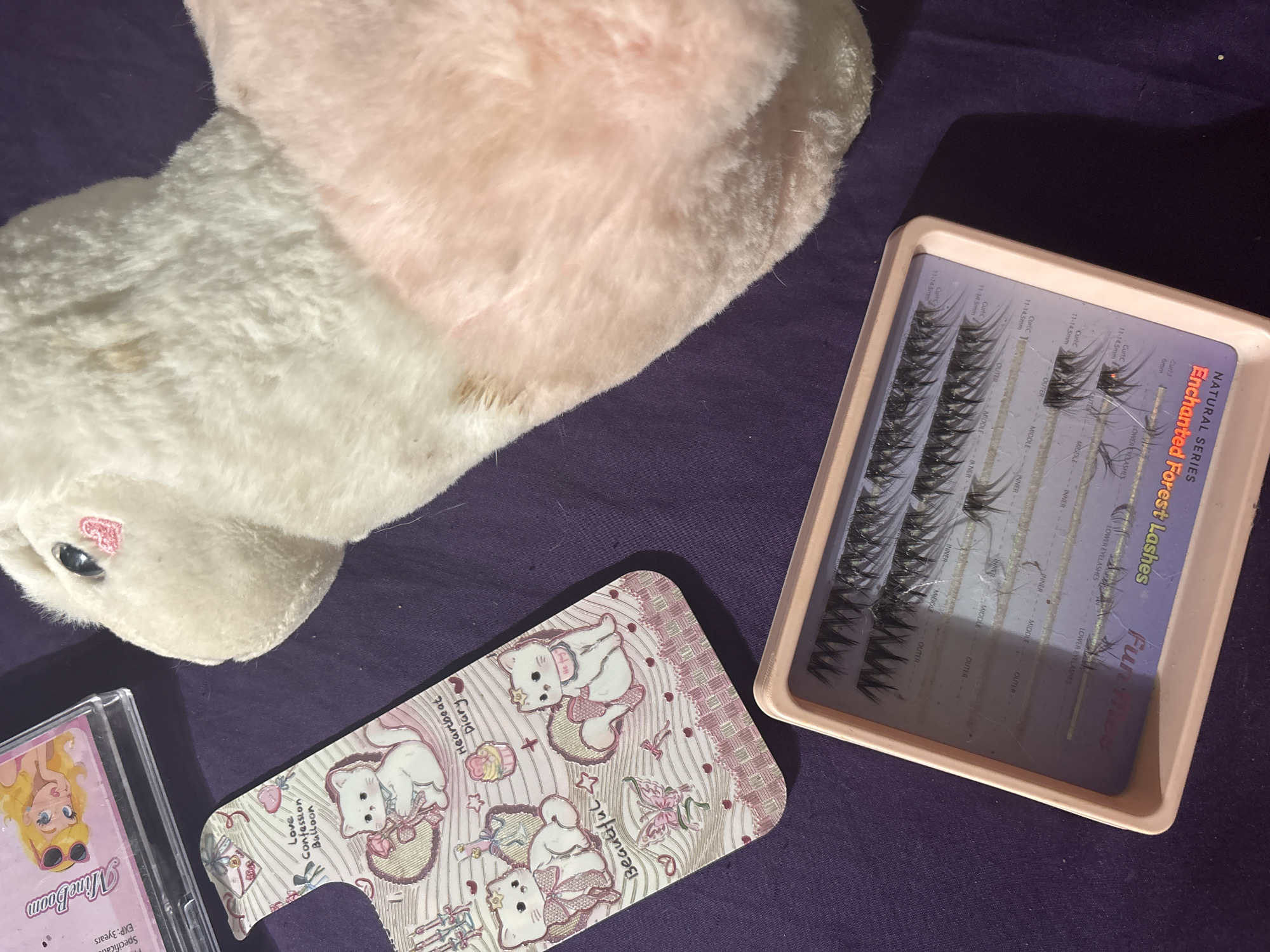 The image displays a collection of items on a dark purple surface. In the foreground, a light pink tray contains several pairs of false eyelashes. The tray has markings indicating different lash placements such as "OUTER," "MIDDLE," and "INNER," as well as "CURVE 11-14.5mm" and "CURVE 8mm." Above the false eyelashes is a pink and white patterned phone case featuring illustrations of cute cats and various decorative elements. To the right of the phone case and partially obscuring the top right corner of the image is a plush toy. The toy is predominantly white with pink accents, resembling a rabbit or a similar soft animal. In the upper left corner, a clear plastic case with a pink lid is visible, containing what appears to be a CD or a similar media format. The lid of this case features an illustration of a cartoon character with blonde hair and sunglasses. The scene is illuminated by overhead lighting, casting shadows on the surface. There is no indication of the location in North Jakarta, Indonesia.