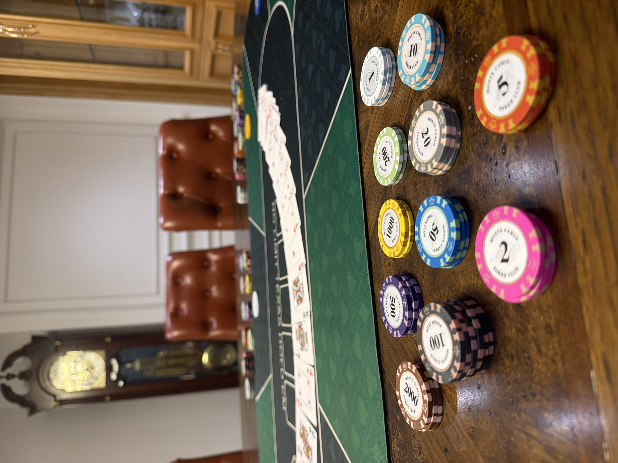 In the city of Xuân Trường - Đà Lạt, Vietnam, a poker game is in progress on a wooden table. A green felt poker mat with various markings is laid out, upon which a fan of playing cards is spread. Surrounding the mat are several stacks of poker chips in various denominations and colors, including 2, 5, 10, 20, 50, 100, 200, 500, and 2000. In the background, two tufted leather chairs are positioned, and a grandfather clock stands to the left, with a wooden cabinet to the right.