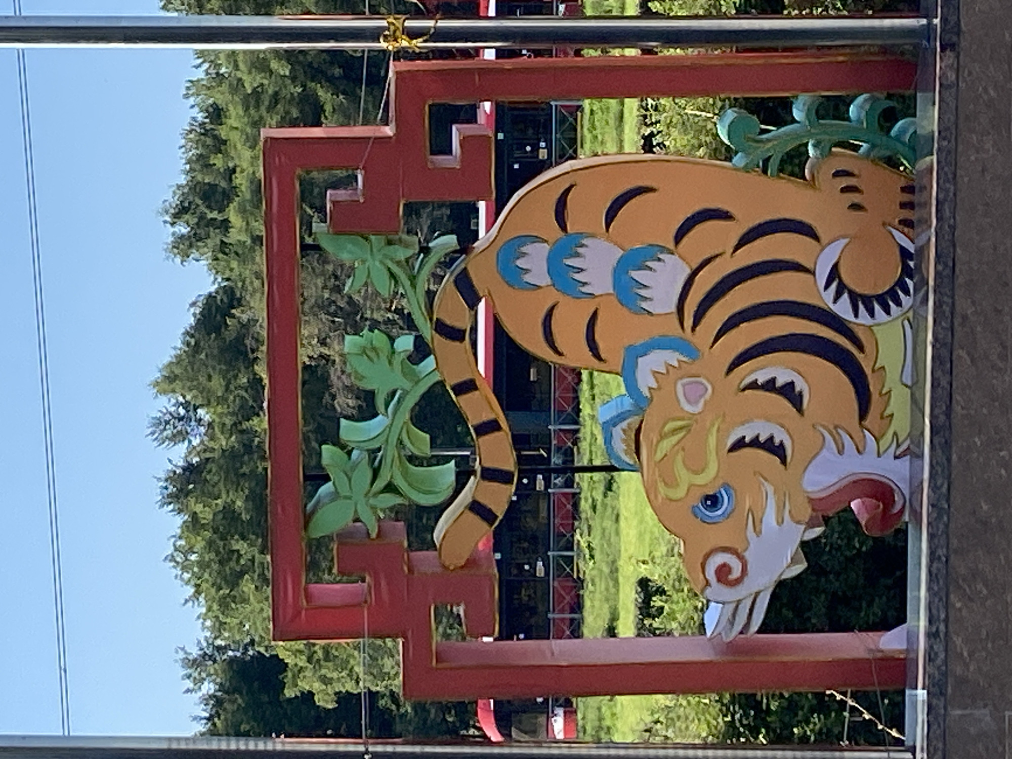 The image shows a colorful, cartoonish tiger decoration against a backdrop of trees and a blue sky. The tiger is predominantly orange with black stripes and blue accents. It is depicted in a playful, arched pose, with its mouth open and tongue visible. Green leaf-like elements and blue, petal-like shapes are incorporated into the design. The tiger is framed by a red, ornate structure with a gold trim. In the background, there is a grassy area with trees and a red bus or tram. The scene is outdoors, under a clear sky, suggesting a public space. The decorative elements and the presence of public transport in the background may indicate an urban setting within Saint Petersburg, Russia.