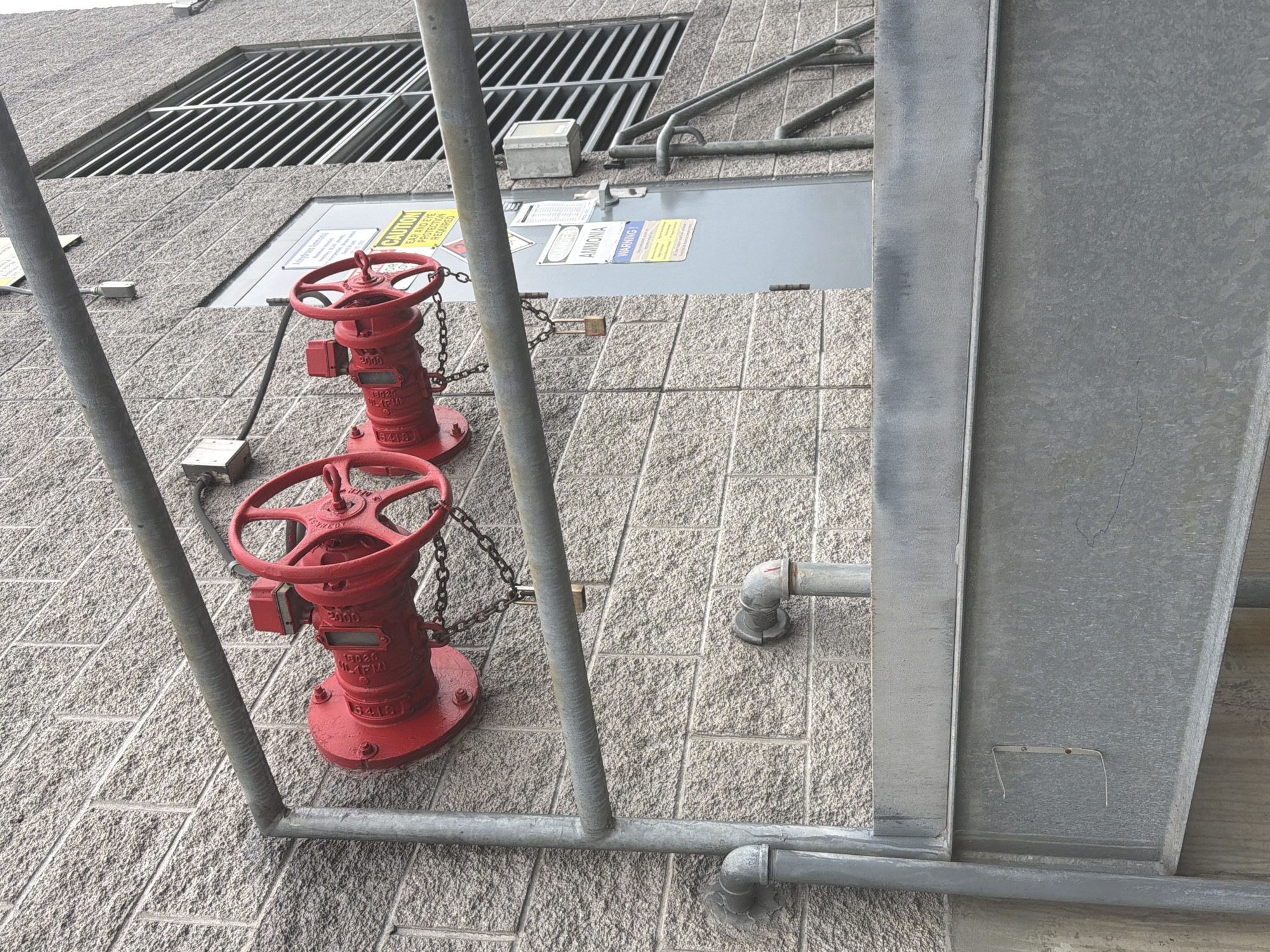 The image depicts a section of an industrial building's exterior wall in Murfreesboro, United States of America. The wall is constructed of light gray, textured brickwork. Two large, red industrial valves with prominent handwheels are mounted to the wall, each secured with chains and padlocks. One valve is labeled with "2000" and what appears to be a manufacturer's name partially obscured. A horizontal galvanized metal pipe runs above the valves, supported by vertical pipes and brackets. To the right of the valves, a gray metal door is visible, adorned with several safety and informational signs. One sign prominently displays "DANGER AMMONIA" and "WARNING." Above the door, a large window or vent with horizontal silver louvers is present. In the foreground, a section of a grey metal structure, possibly stairs or a platform, occupies the lower portion of the frame. The environment appears to be an outdoor industrial setting, with no people visible in the frame.