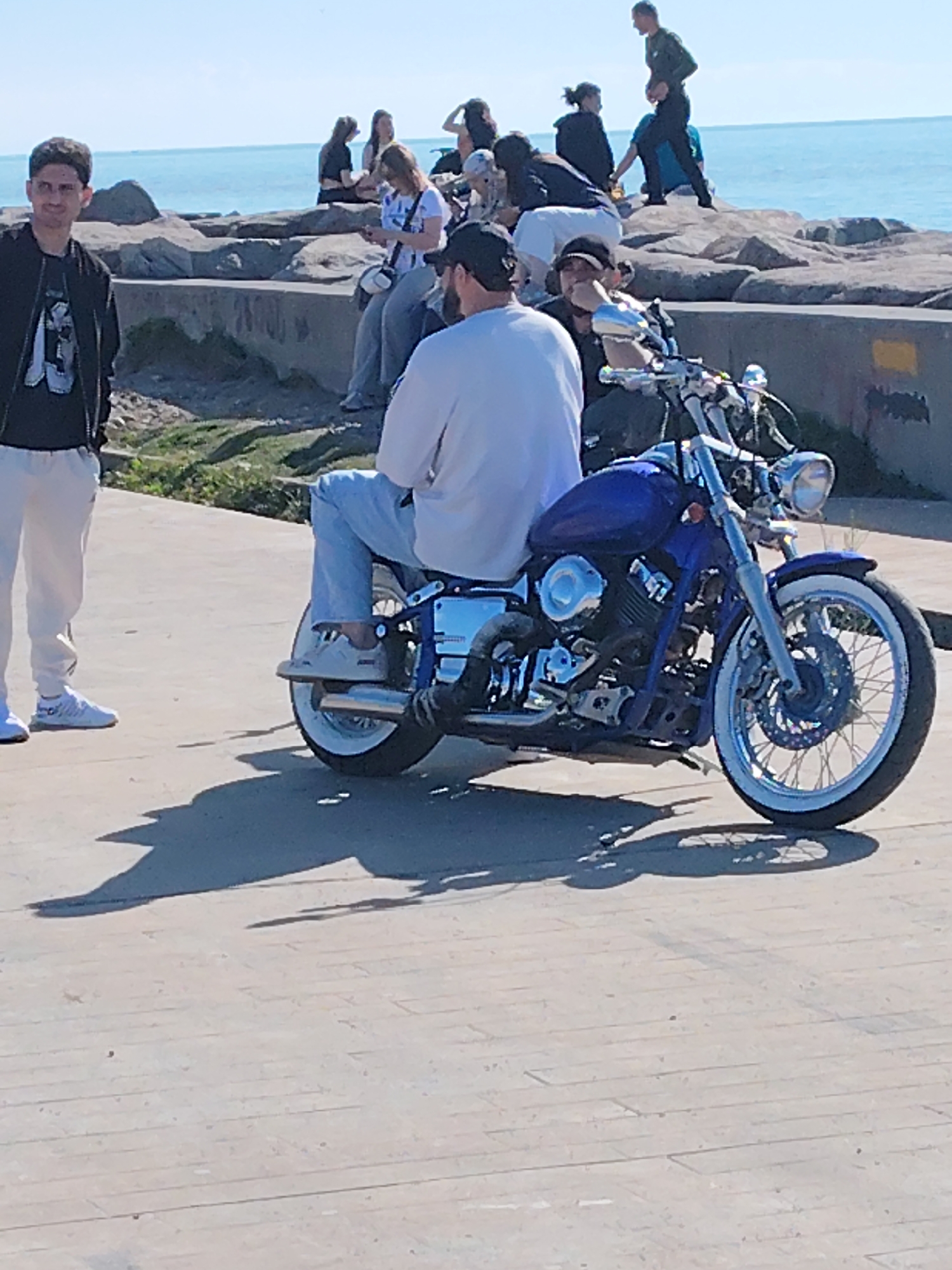 The photograph captures a scene in Batumi, Georgia, near the coastline. A blue motorcycle is the prominent subject, parked on a concrete surface alongside a pedestrian walkway. A man sits on the motorcycle. Another man stands near the bike. In the background, a group of people is gathered along a rocky shoreline overlooking the sea. The day appears sunny and bright.