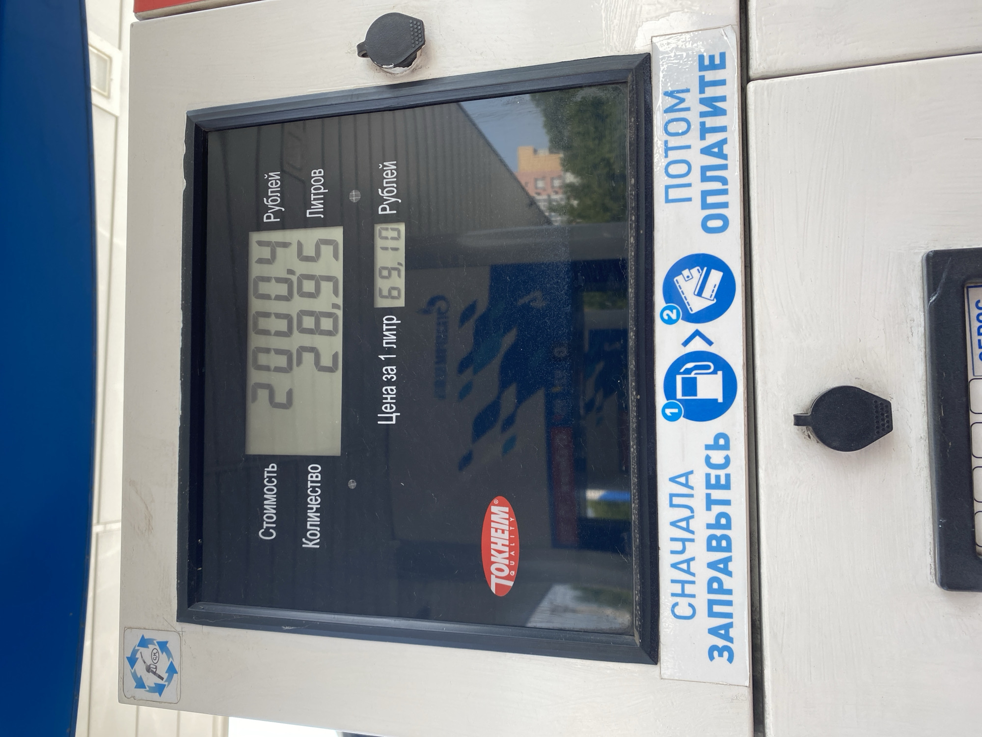 The image shows a gas pump display at a gas station in Novokuznetsk, Russia. The display shows the cost of fuel at 2000.4 rubles, the volume dispensed at 28.95 liters, and the price per liter at 69.10 rubles. There is a Tokheim Quality logo on the pump. Below the display, there are instructions in Russian that read "First, refuel, then pay." The scene is well-lit and appears to be during the daytime. There are no people visible in the image.