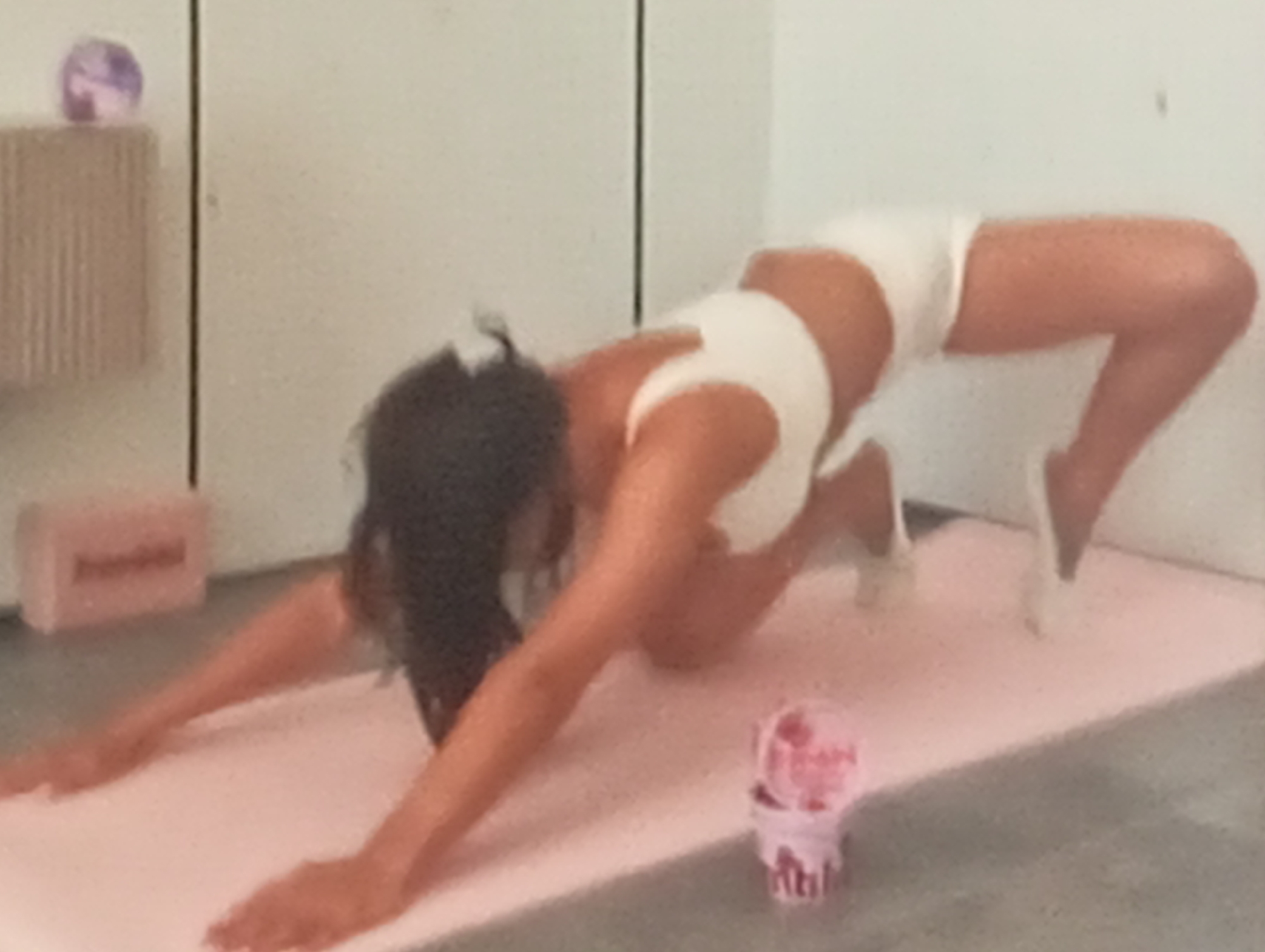 A woman is performing a yoga pose on a pink yoga mat. She is in a bridge pose with her back arched and her hands extended on the mat in front of her. She is wearing white athletic shorts and a matching sports bra. Her dark hair is falling forward. In the background, there is a light-colored wall and on the left side, a textured object with a small purple sphere on top. A pink yoga block is visible on the floor to the left of the mat. To the right of the woman on the mat, there is a small pink container with white text. The lighting suggests it is daytime. The scene appears to be indoors, likely a home or studio space, given the yoga mat and equipment. The woman's posture and the presence of yoga props suggest a focus on physical activity and well-being.
