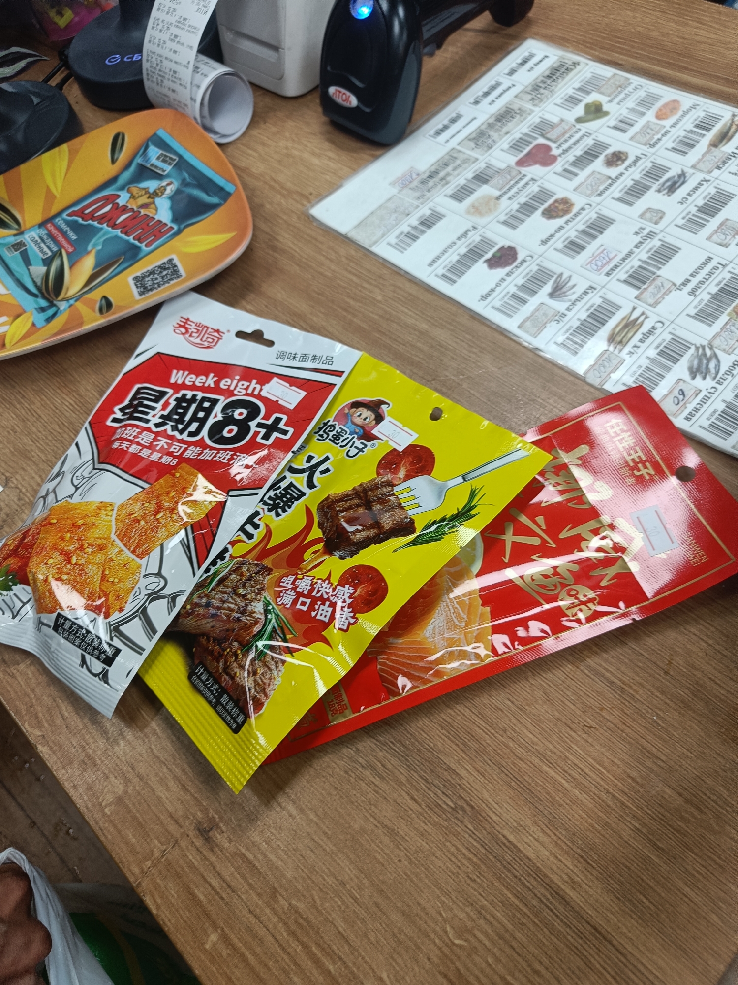 The image shows a close-up of a wooden table with various items. In the foreground, there are three packages of snacks, two of which are brightly colored. One is yellow with images of grilled meat and a fork, and the other is red with Chinese characters. The third package is white and red, also with Chinese characters, and features the text "Week eight" and "星期8+". To the left of these packages is a decorative plate featuring a packet of sunflower seeds labeled "ДЖОННИ" in Russian, along with the words "семечки" (sunflower seeds), "качественной" (high-quality), "обжарки" (roasting), and "солёные" (salted).
In the background, a retail or checkout counter is visible, with a black scanner-like device, a white receipt printer, and a roll of receipts. To the right of these is a sheet of paper covered in small squares, each containing an image of food or an item and what appears to be a barcode and price in Russian. Some visible text on these squares includes "Толстолоб" (carp), "юкола вяд." (dried fish), "Вобла сушеная" (dried roach), "Сайра х/к" (mackerel), "Иваси" (sardines), "Хамса с/с" (anchovies), "Щука ломтики" (pike slices), "Килька п/с" (sprat), "Рыба соленая" (salted fish), "Капуста" (cabbage), "Салаты по-кор." (salads), and "Грибы маринов." (marinated mushrooms). The overall impression is that of a retail environment, possibly a convenience store or a supermarket, with products laid out on a table. The presence of both Russian and Chinese text suggests a location where these cultures might intersect, possibly in a port city or an area with significant trade. The lighting suggests an indoor setting during the day.