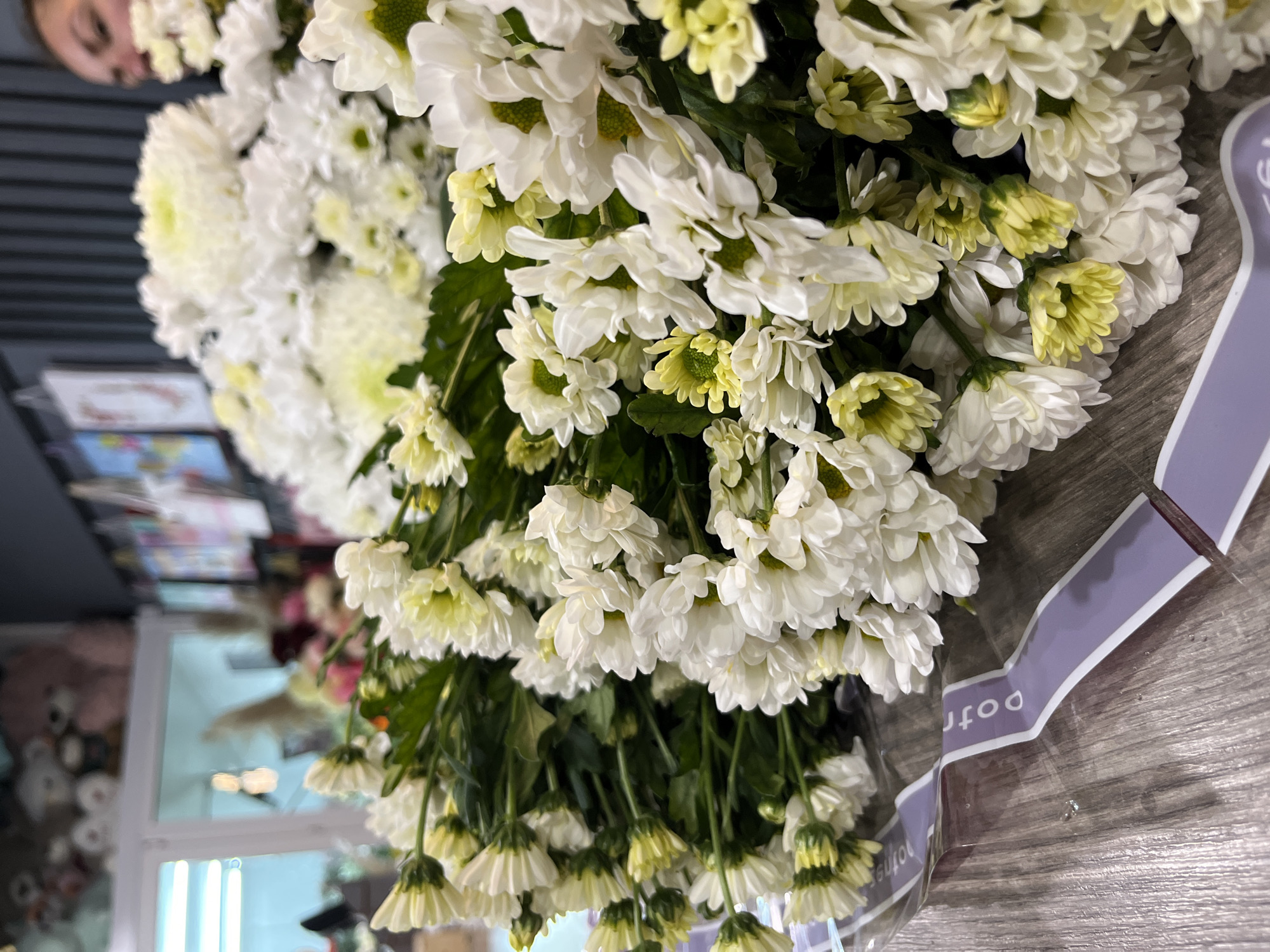 A close-up, high-angle shot captures a large bouquet of white chrysanthemums, some with light green centers. The bouquet is wrapped in clear plastic and tied with a lilac-colored ribbon that says "potri". The background appears to be a store setting with blurry objects and reflections suggesting a commercial space. Human reflection is seen in the left upper corner. The photo was taken in the city of Klin, Russia.