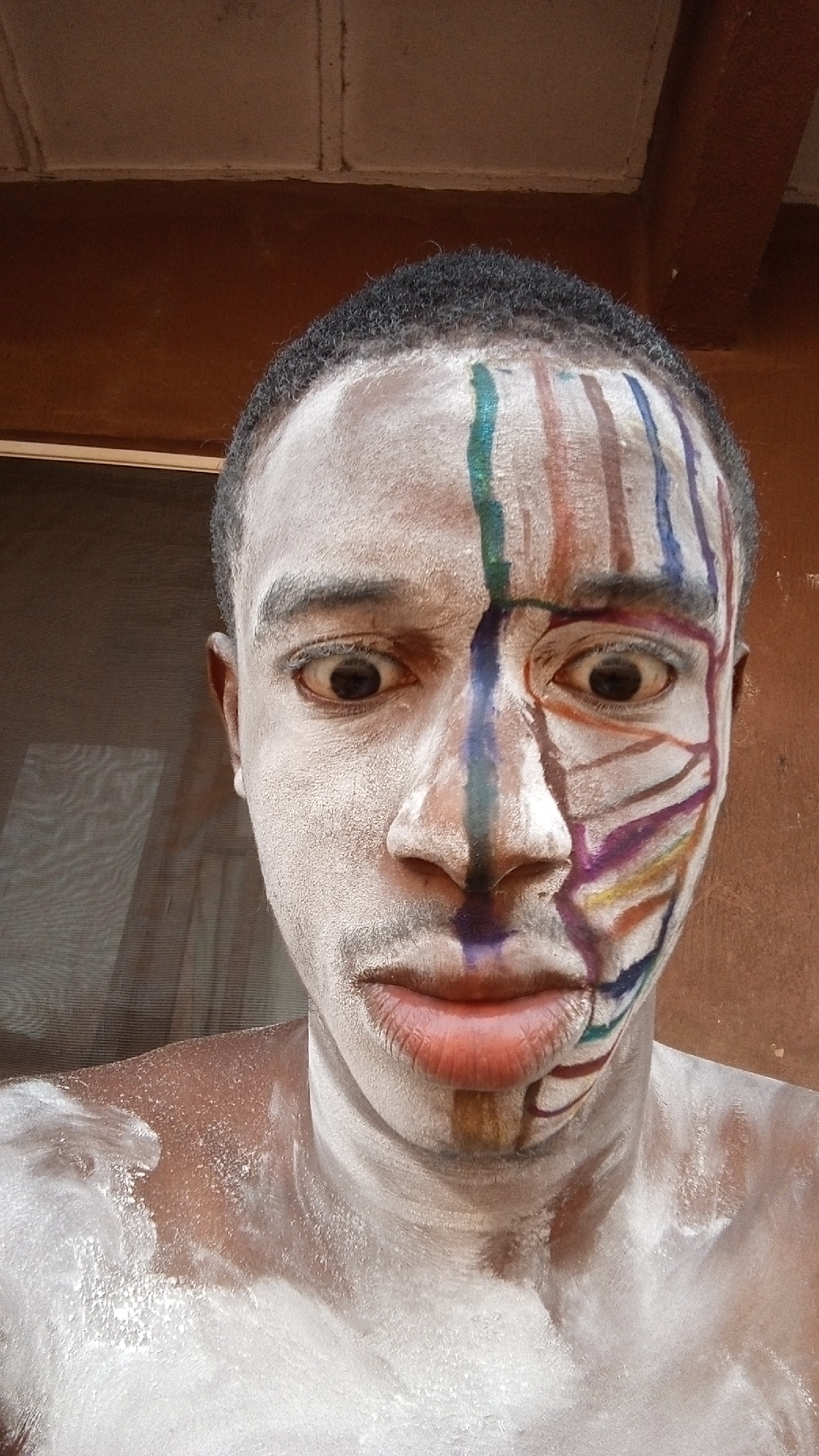 A young, dark-skinned individual with short, cropped hair is the main subject of the image. The individual's face is partially covered in white powder, with colorful stripes of paint on one side, running down from the forehead. The paint colors include green, brown, and blue. The background suggests an interior setting with a brown wall and a section of what appears to be a window. The location is Unknown, Unknown.