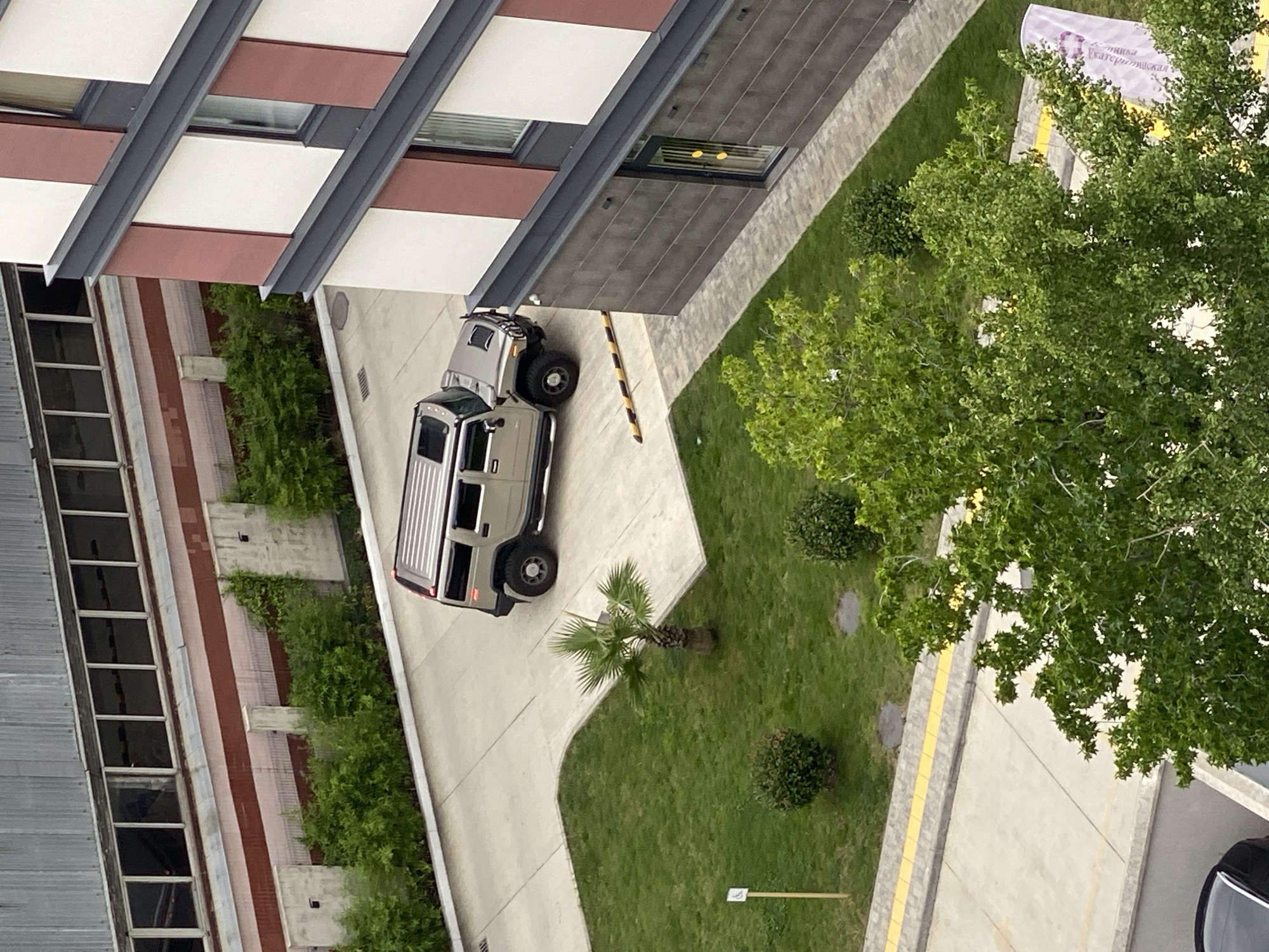 An elevated, high-angle, medium shot captures a beige Hummer H2 parked on a paved area adjacent to a modern building with reddish-brown and white facade elements and a dark grey base. The Hummer is positioned in the middle of the frame, with its front facing right. To the left of the Hummer, a small palm tree stands on a grassy area. In the foreground, a large green tree with dense foliage partially obscures the lower portion of the image. The background features a long, grey industrial-style building with numerous windows. The scene appears to be an urban environment, possibly a residential complex or a commercial area. The time of day is unclear due to overcast lighting. There are no people visible in the image.