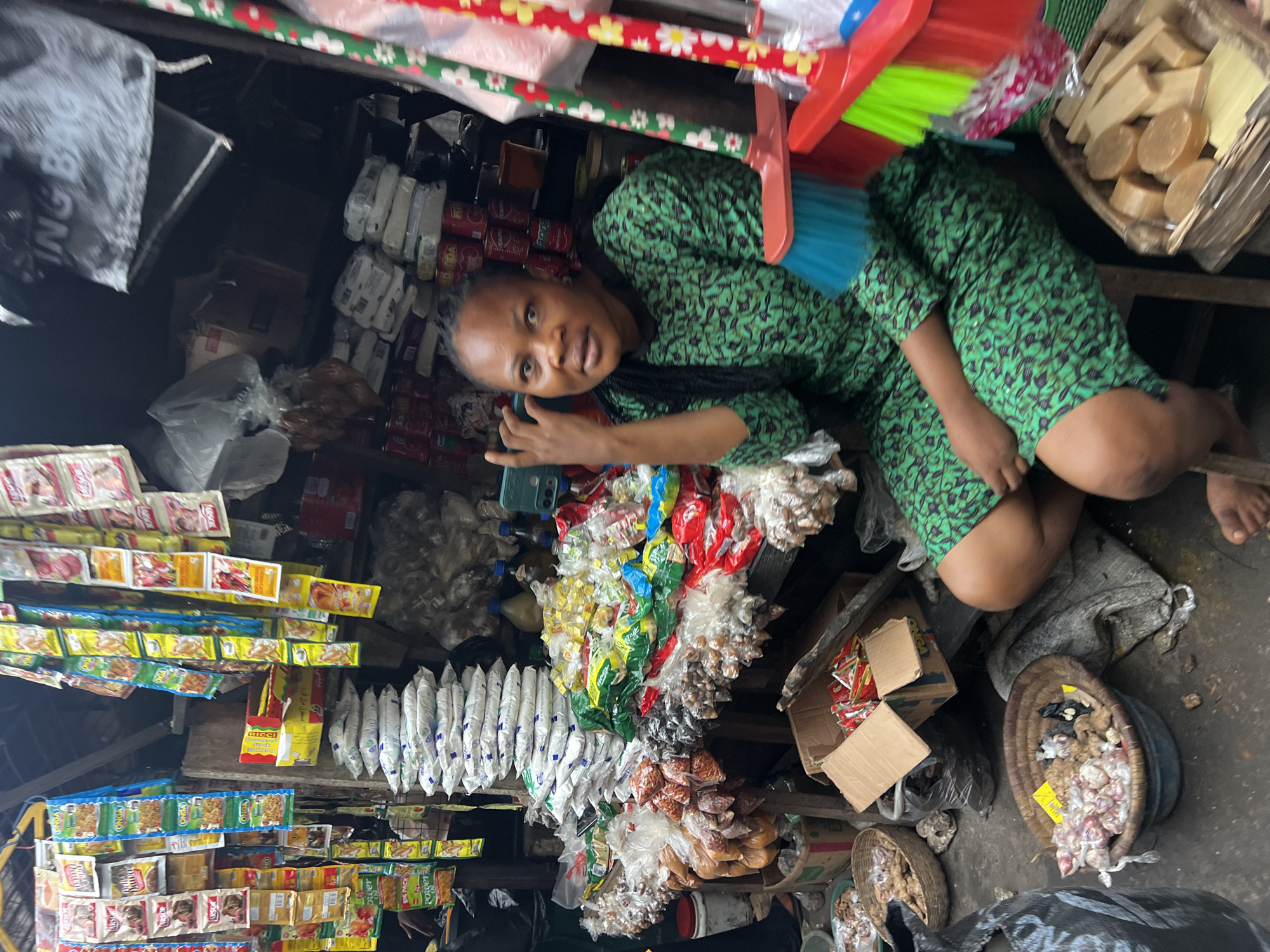 A medium shot shows a woman in a green patterned dress sitting in an outdoor market stall in Nkpor, Nigeria. She is looking towards the camera with her right hand raised, fingers splayed, as if gesturing. The stall is filled with various goods, including bags of spices and produce, stacked white bags, and packets of seasonings hanging from the ceiling. In the foreground, baskets of garlic and what appear to be bars of soap are visible. A broom with a red and green handle is also present. The ground is dirt and concrete, suggesting an open-air market. The overall impression is one of a bustling marketplace.