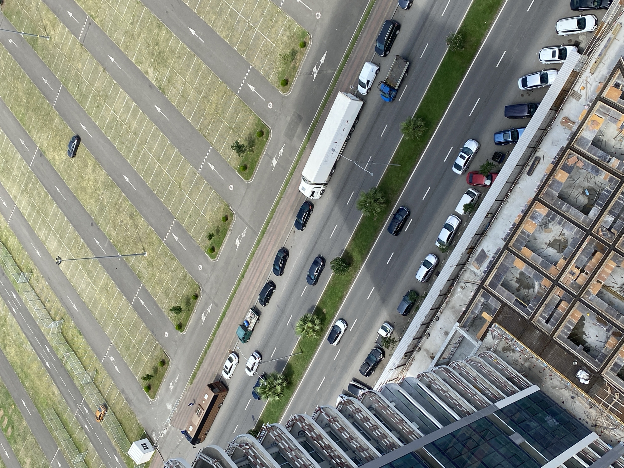 An aerial, high-angle, daytime, establishing shot captures an urban scene in Batumi, Georgia, featuring a large parking lot and a busy multi-lane road bordered by a modern building under construction and a completed high-rise residential building. The parking lot is vast and mostly empty, with rows of marked parking spaces delineated by white lines on asphalt and grass. A few vehicles are visible within the parking lot, including an orange car, a dark-colored sedan, and a white boxy structure resembling a small building or kiosk.
The main focus of the image is the multi-lane road where traffic is flowing. Numerous vehicles, including cars, a bus, a large white semi-trailer truck, and a small blue flatbed truck, are present on the road. Several cars are parked along the side of the road in designated pull-off areas or parking bays. The road is lined with green grass medians and some palm trees.
To the left of the road is a completed, modern, multi-story residential building with balconies and glass-paneled windows. Adjacent to this building and extending further into the foreground is a construction site. This area displays exposed concrete foundations, rebar, wooden formwork, and stacks of building materials, indicating ongoing construction work. A grey metal fence separates some sections of the construction site and roadside. The overall environment appears to be a developed urban area with a mix of infrastructure, residential, and commercial elements.