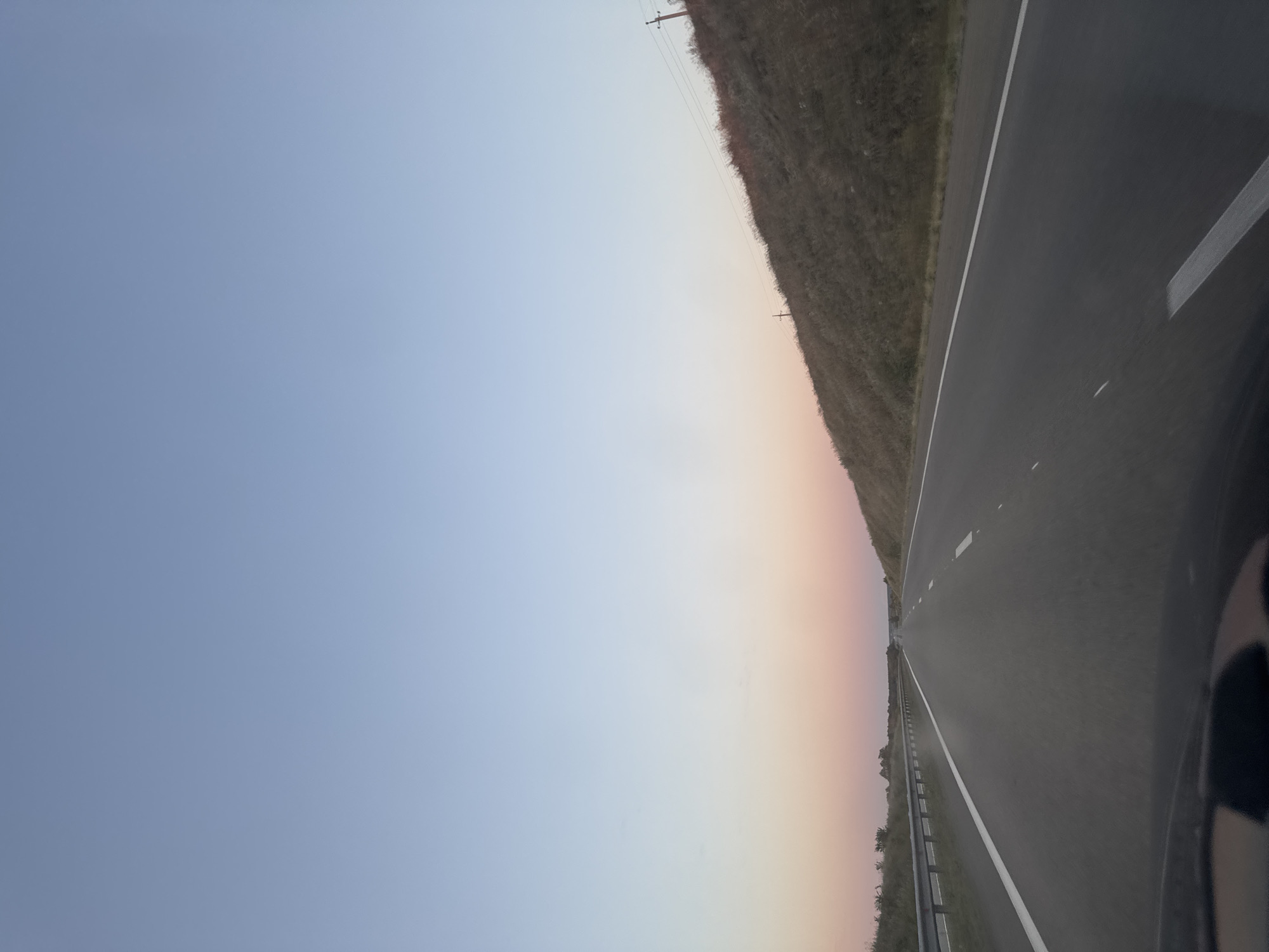 The image depicts a highway in Pridorozhnyi, Russia, during what appears to be either sunrise or sunset, as indicated by the gradient of pastel colors in the sky, transitioning from a light blue at the top to hues of pink and orange near the horizon. The road itself is asphalt and shows white lane markings. On the left side of the road, there is a metal guardrail. To the right of the road, a grassy embankment rises, sparsely dotted with vegetation and a utility pole with wires. The perspective is from within a vehicle, looking forward down the highway, with the lower part of the image showing the dashboard and windshield area. The overall environment is rural and open. There are no people or animals visible in the frame.