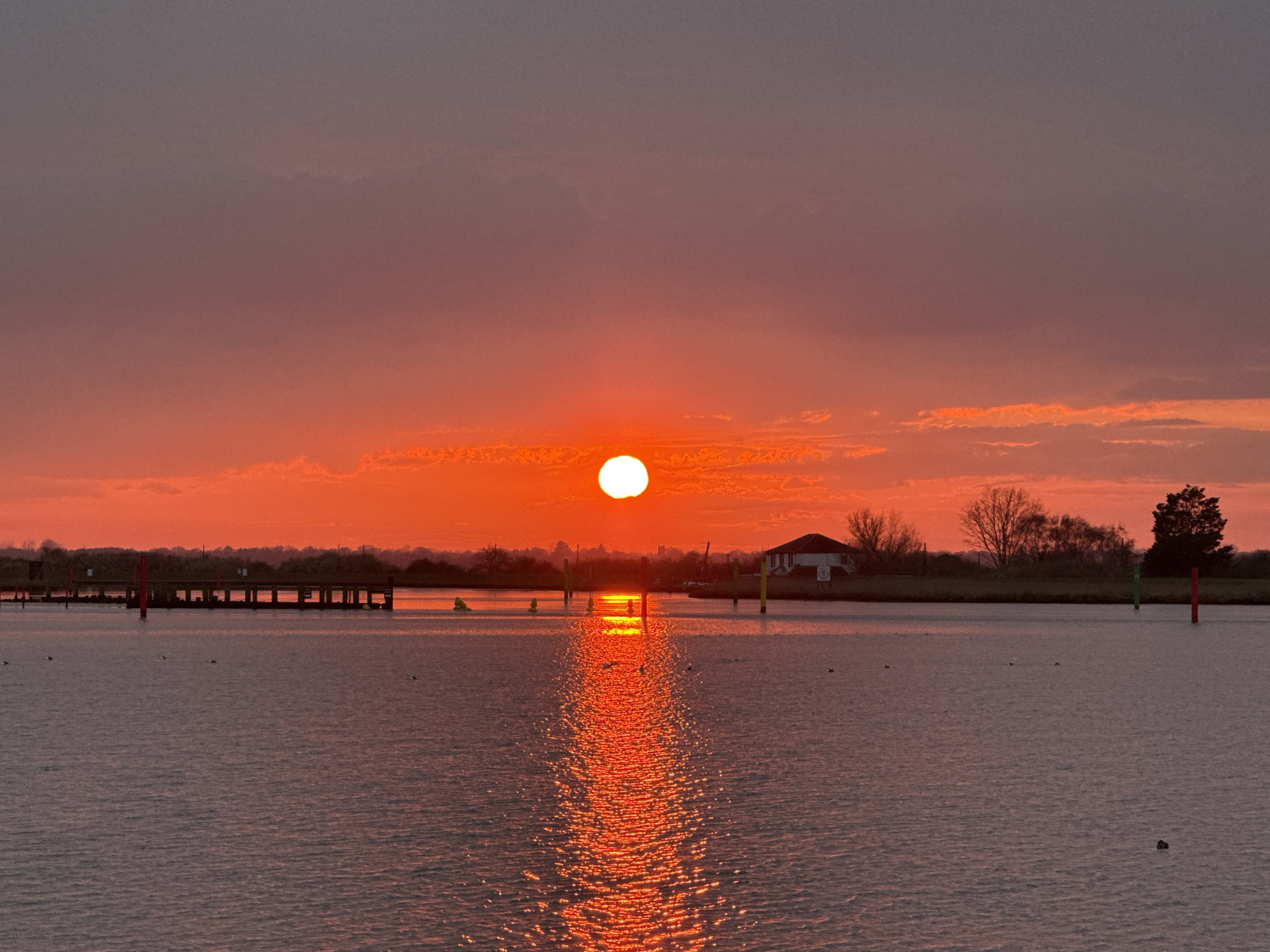 A wide-angle shot shows a red sunset over water. The sun is positioned centrally and reflects on the water. Some dark, low clouds are visible above the sun. A small pier is visible on the left. There is a white house on the right side of the photograph, with a sign on it, as well as a number of trees to the right of the house. Several posts are in the water, with different colored tops. A small boat can be seen floating. This is likely Breydon Water, a tidal estuary located in Great Yarmouth, United Kingdom.