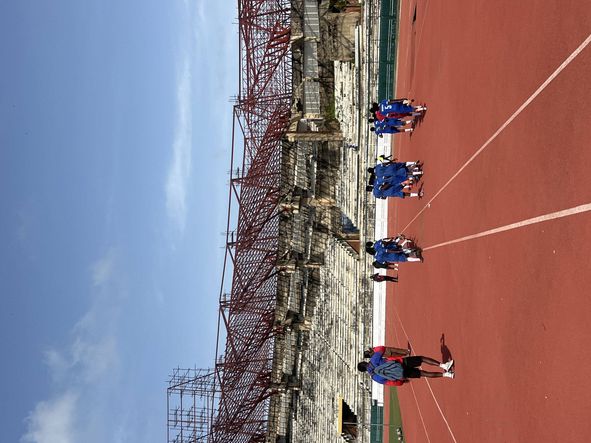 In New Stadium, Nigeria, a group of young men in blue soccer uniforms are walking across a red running track. Some wear cleats, while others have on sneakers. A person in a red backpack and black shorts walks away from the camera on the track. In the background, an old, partially constructed stadium with exposed red metal beams and concrete seating stands under a bright blue sky with scattered clouds.