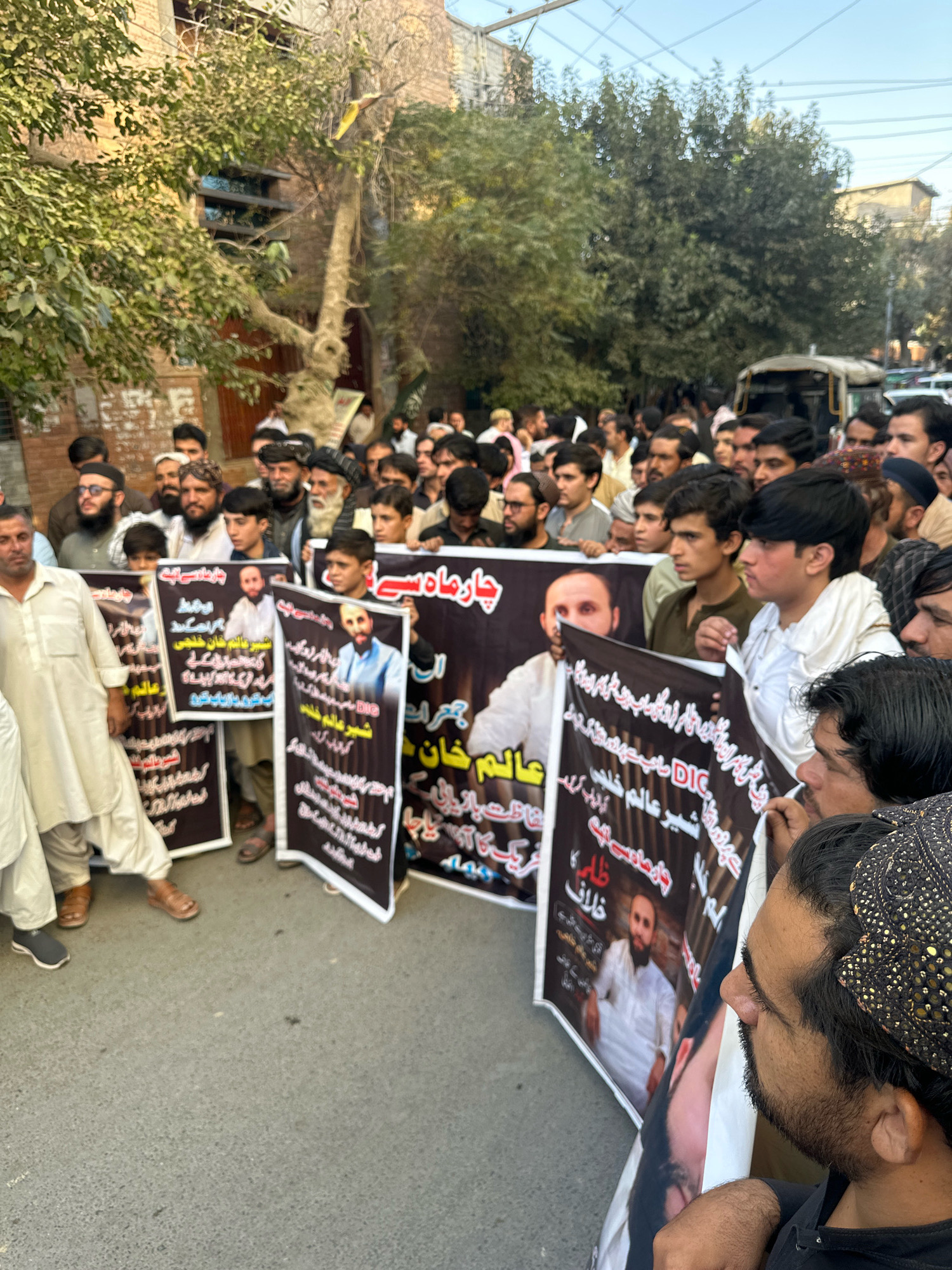 A group of men and boys are gathered in an outdoor setting, possibly a street in Quetta City Tehsil, Pakistan. They are holding up several large banners, some of which feature portraits of individuals and text in an unfamiliar script. The people in the image appear to be looking forward, and some are holding the banners. The environment includes trees, buildings in the background, and a clear sky. The overall scene suggests a protest or demonstration.