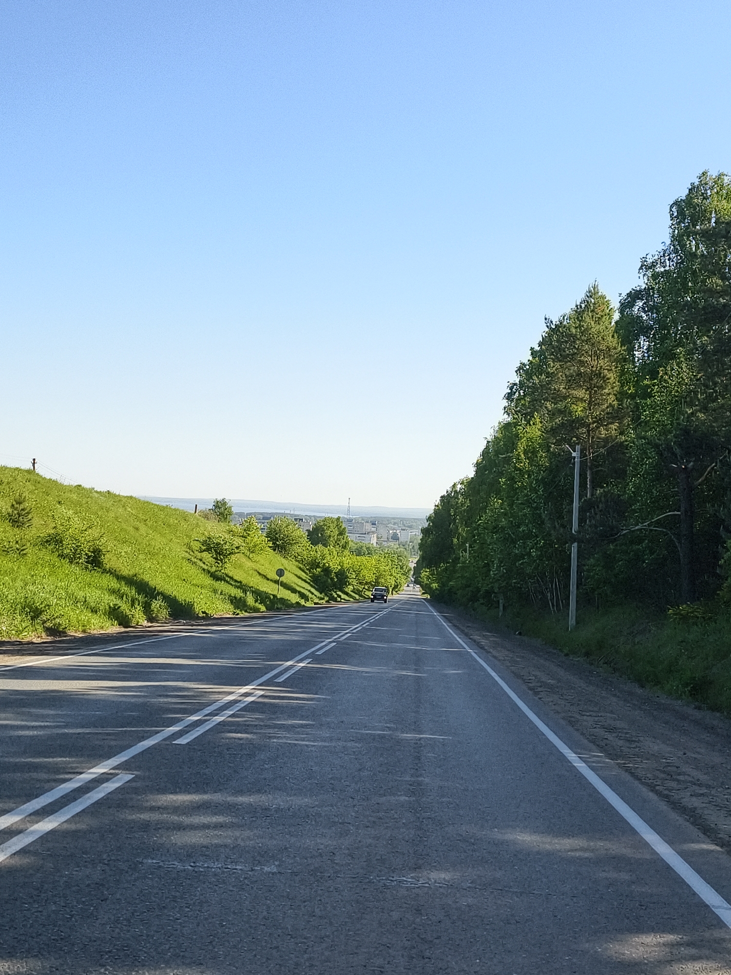 The image depicts a straight asphalt road leading towards a distant cityscape under a clear blue sky. The road is marked with white lines, indicating traffic lanes. Lush green vegetation covers the embankment to the left of the road, while a dense line of trees borders the road on the right. A single car is visible in the distance on the road. The location is identified as Chaikovsky, Russia, based on the visual elements and the known geography.