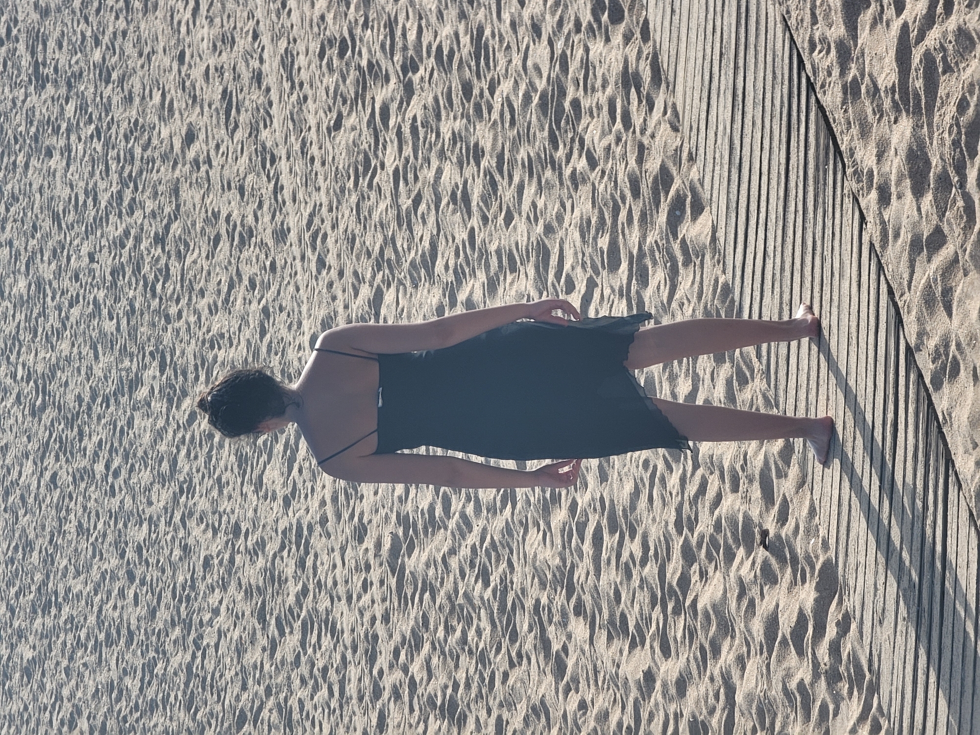 This media file shows a person lying on a sandy beach, with their head towards the top of the frame. They are wearing a dark-colored dress with thin straps. The person's body extends diagonally across the frame, with their feet towards the bottom right. The sand has a wavy texture, suggesting it has been disturbed by wind or water. To the right of the sand, a wooden boardwalk runs parallel to the edge of the frame. The lighting suggests it is daytime, with shadows cast by the boardwalk onto the sand. The scene appears to be a beach or coastal area, given the presence of sand and a boardwalk. The orientation of the image is landscape. The person's pose and the serene setting evoke a sense of relaxation or repose. There is no visible text in the image.