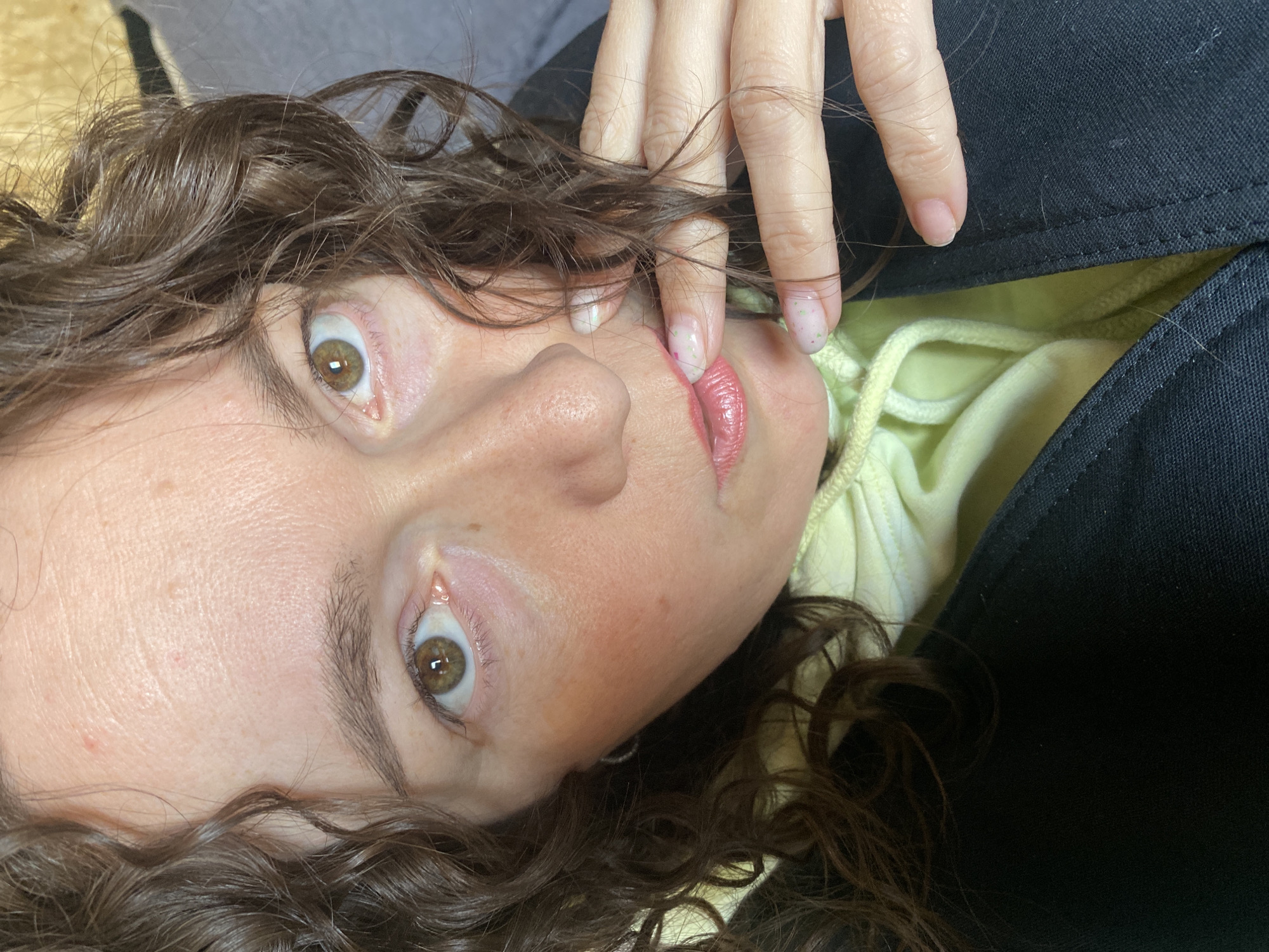 A woman with brown curly hair and hazel eyes is lying down, facing upwards. Her right hand is near her face, with fingers slightly touching her lips. She is wearing a light yellow hoodie underneath a black jacket. The image appears to be taken at a close-up, slightly angled perspective. There is no identifiable indication of being located in Ol'khovskoye, Russia.
