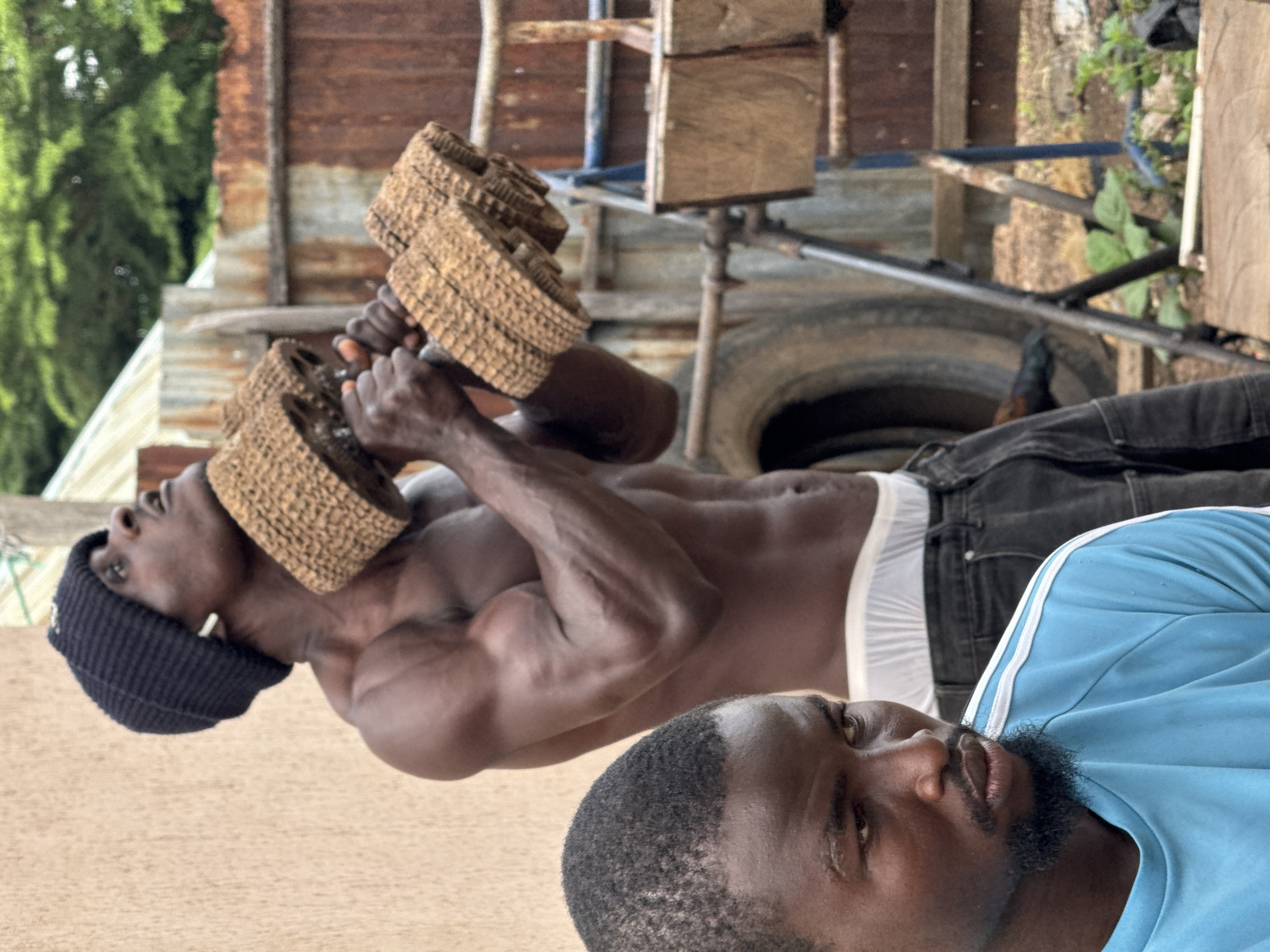The image captures a scene likely outdoors, possibly in a workout area, with two men prominently featured. The foreground is dominated by the head and upper torso of a man wearing a light blue t-shirt with white stripes on the shoulders. He has a dark complexion, short black hair, and a beard. He is looking downwards and slightly to his left, with a thoughtful or concerned expression.
In the mid-ground and background, another man, also with dark skin, is engaged in weightlifting. He is shirtless, displaying a muscular physique. He wears a black beanie and white earbuds. He is holding up two large, textured dumbbells, appearing to be made from woven material, possibly straw or rough fibers, with wooden handles. His head is tilted upwards as he performs his exercise.
The environment appears somewhat rugged and utilitarian. Behind the men, there's a textured, light-colored wall on the left, and to the right, a rusty corrugated metal fence. A large tire is visible in the background, along with some metal scaffolding and wooden structures. Green foliage and trees can be seen in the upper right portion of the image, suggesting an outdoor setting. The overall impression is one of an informal, perhaps makeshift, outdoor gym. The image has a slightly grainy texture.
While the image itself does not explicitly state the location, the prompt specifies the city of Akwanga, Nigeria. This context suggests that the scene is situated within this Nigerian city, likely in a less developed or more rustic part of the urban environment, possibly a residential area or a communal space adapted for physical training.