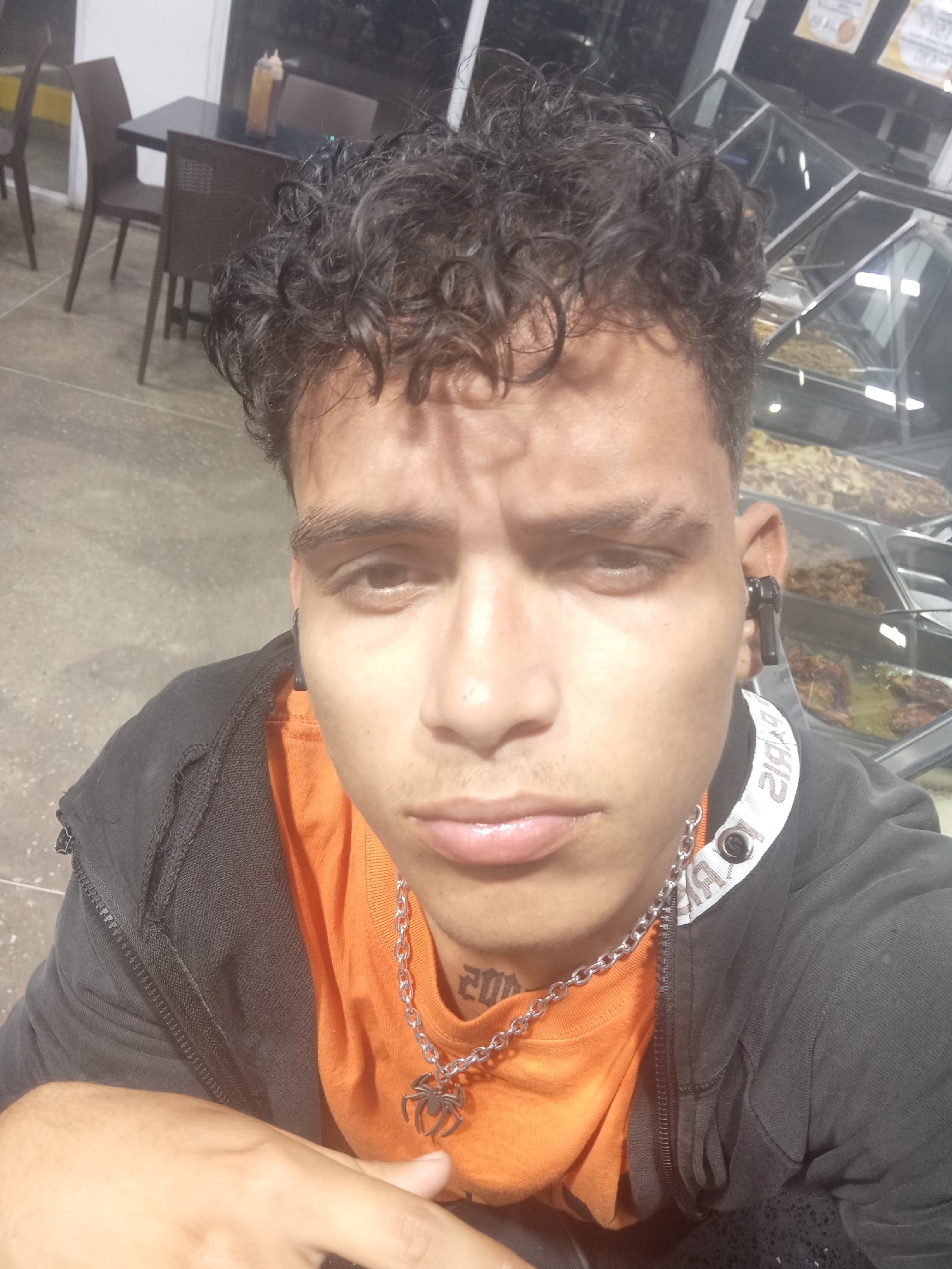 A close-up shot of a young adult male with curly hair and dark skin is shown. The male is wearing a dark hoodie over an orange t-shirt, and a silver chain with a spider pendant. The tattoo "2012" is visible on his neck. He has a wireless earbud in his right ear.
Behind him, partially visible, is a food display case with prepared food items. In the background, there is a table with chairs. The scene appears to be indoors, possibly a food establishment, or a small restaurant, in Juan Griego, Venezuela.