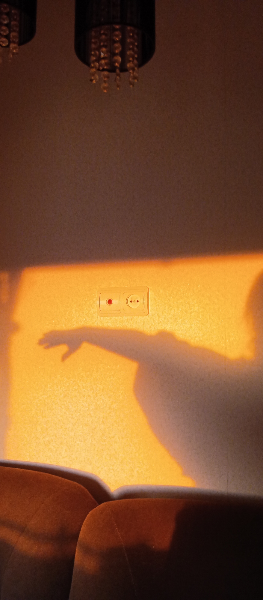 A vertically-oriented photograph captures an interior scene, exhibiting a wall with a shadow cast upon it, and a section of an upholstered chair at the bottom. The wall is light in color and textured. A European-style electrical outlet is installed on the wall, featuring a red button on the left receptacle. A shadow of an arm and hand is projected onto the wall, appearing to reach toward the outlet. Above the outlet, the wall darkens. The upper portion of two ornate chandeliers is visible at the top of the frame. The light source illuminating the shadow seems to be from the left, casting a warm glow on the wall. The bottom of the image is occupied by the top of a chair, which is upholstered in a brown fabric. The setting appears to be indoors, possibly in a home or office space. Location: Unknown, Unknown.