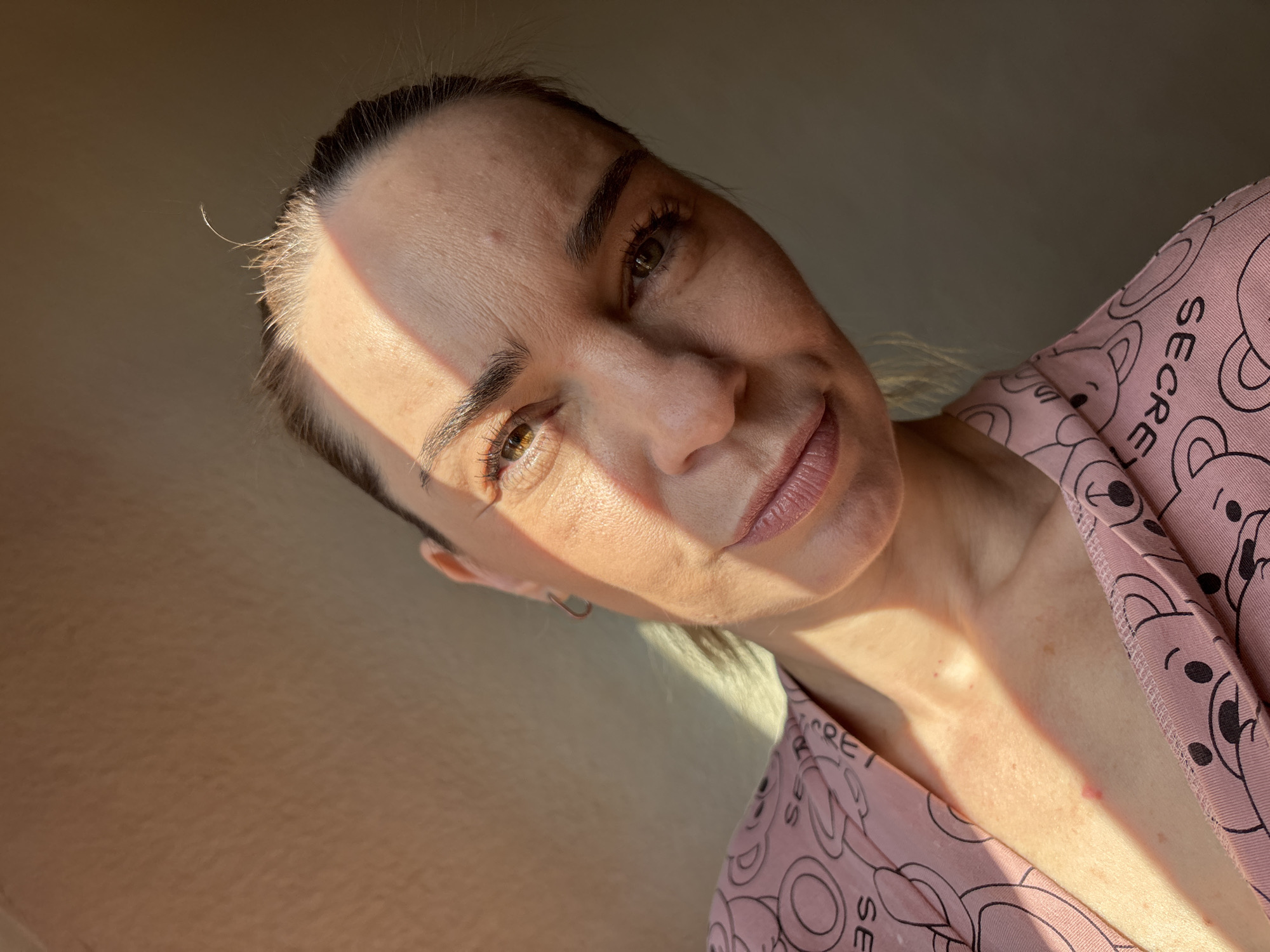 A close-up, high-angle shot captures a woman with light brown hair tied back, facing the camera. A diagonal shadow from sunlight cuts across her forehead and cheek. She wears a pink garment with a repeating pattern of teddy bear faces and the word "SECRET." Her skin shows some redness and light freckling on her chest and neck. The background is a neutral, textured wall. The image is taken indoors, likely in a domestic setting.