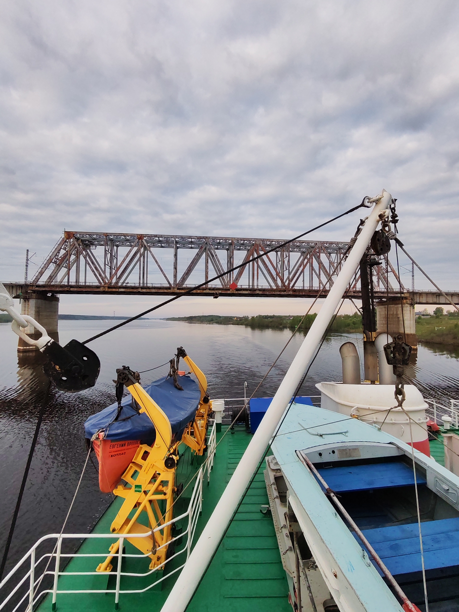 View from a vessel moving on a body of water with a railway bridge in the background. The bridge is a metal truss structure with brown and red coloring, supported by concrete piers. On the vessel's deck, there is an orange lifeboat covered with a blue tarp and a blue rowboat. The deck is green. A white pole and ropes are part of the vessel's equipment. The water is calm with some ripples. The sky is overcast with gray clouds. The location is Tarkanovo, Russia.