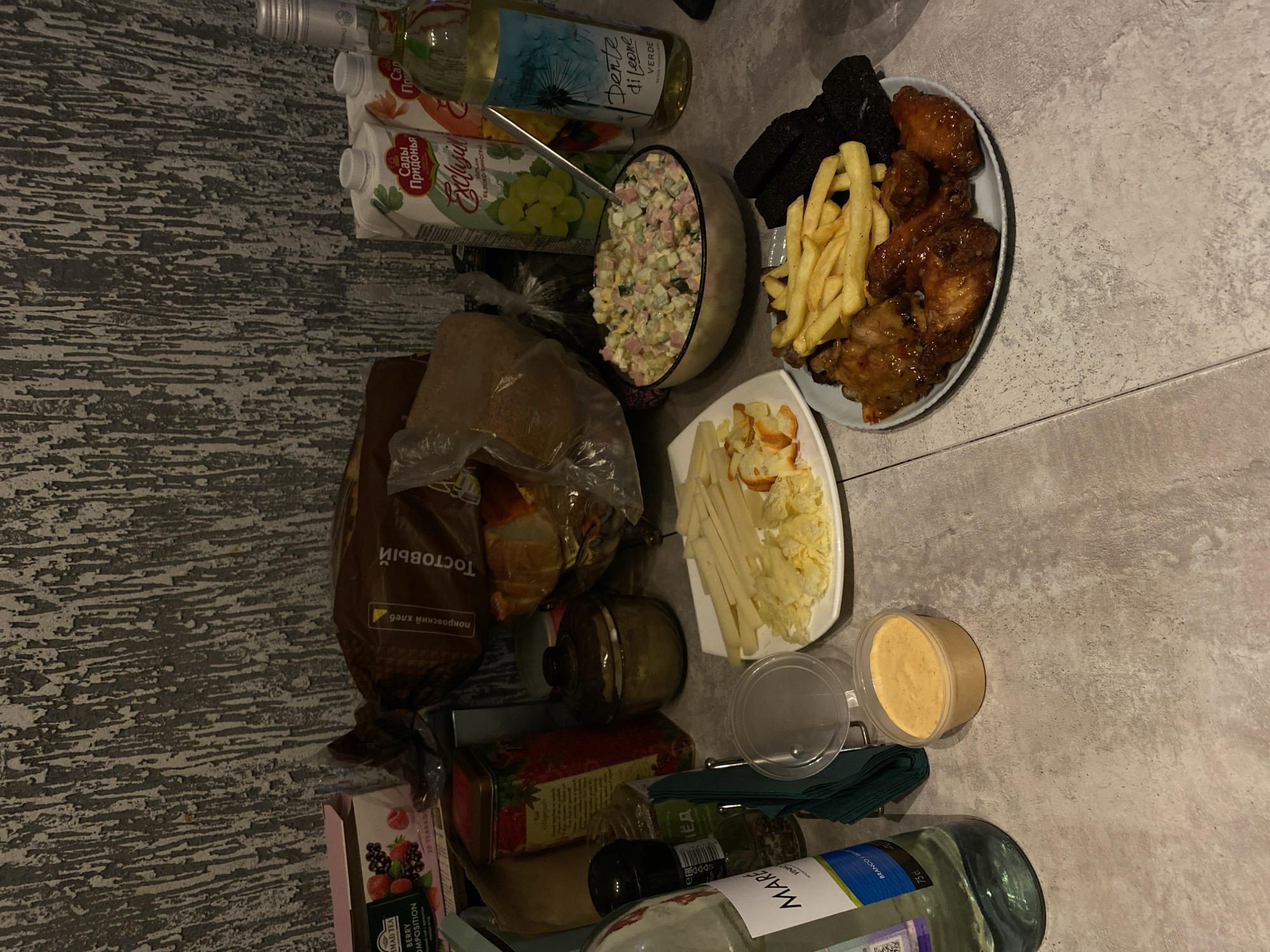 This image captures a casual meal spread laid out on a light gray, textured surface, likely a kitchen counter or table, in Chaikovsky, Russia. The scene is illuminated by artificial indoor lighting, suggesting it's evening.
The table is laden with various food and drink items. Prominently featured is a plate holding golden french fries, several glazed or sauced chicken wings, and dark bread rusks (likely rye bread croutons). Another bowl contains a large portion of what appears to be Olivier salad, a traditional Russian dish with diced vegetables, meat, and mayonnaise. A white rectangular plate displays sliced cheese or possibly potato sticks, along with some lightly fried bread pieces. A small plastic container holds a creamy orange dipping sauce.
Beverages include a bottle of "Dente di Leone VERDE" white wine and a carton of "Сады Придонья" (Sady Pridonya) "ЯБЛОКО ВИНОГРАД" (Apple Grape) juice. Another bottle, labeled "MARE BRANCO", is also visible. A loaf of "Тостовый" (Toast) bread, specifically "покровский хлеб" (Pokrovsky bread), is wrapped in plastic. A box of "MAD TEA BERRY COMPOSITION" tea bags is also present, alongside other unidentifiable jars and bottles. The setup suggests a relaxed gathering or family meal, offering a variety of snacks and dishes.