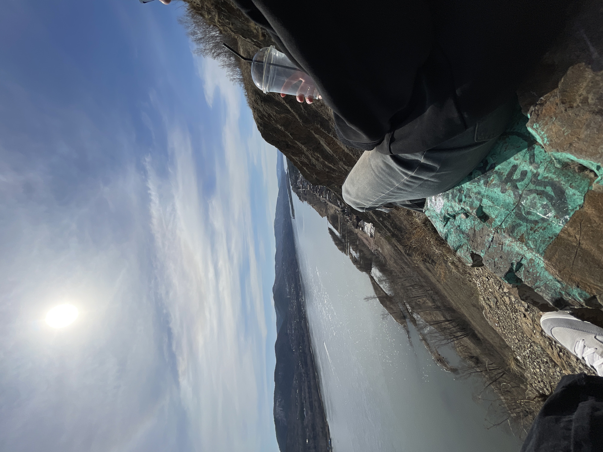 The image depicts a person sitting on a cliff overlooking a wide river. The person is holding a clear plastic cup with a straw. The cliff face is rocky with patches of green and possible graffiti. The river stretches into the distance, flanked by tree-covered mountains. The sky is blue with scattered clouds, and the sun is shining brightly. The person is wearing grey pants and a black jacket. A white shoe can also be seen in the corner. The location is likely in a mountainous area with a large river, and it is possible the city is Krasnoyarsk, Russia.