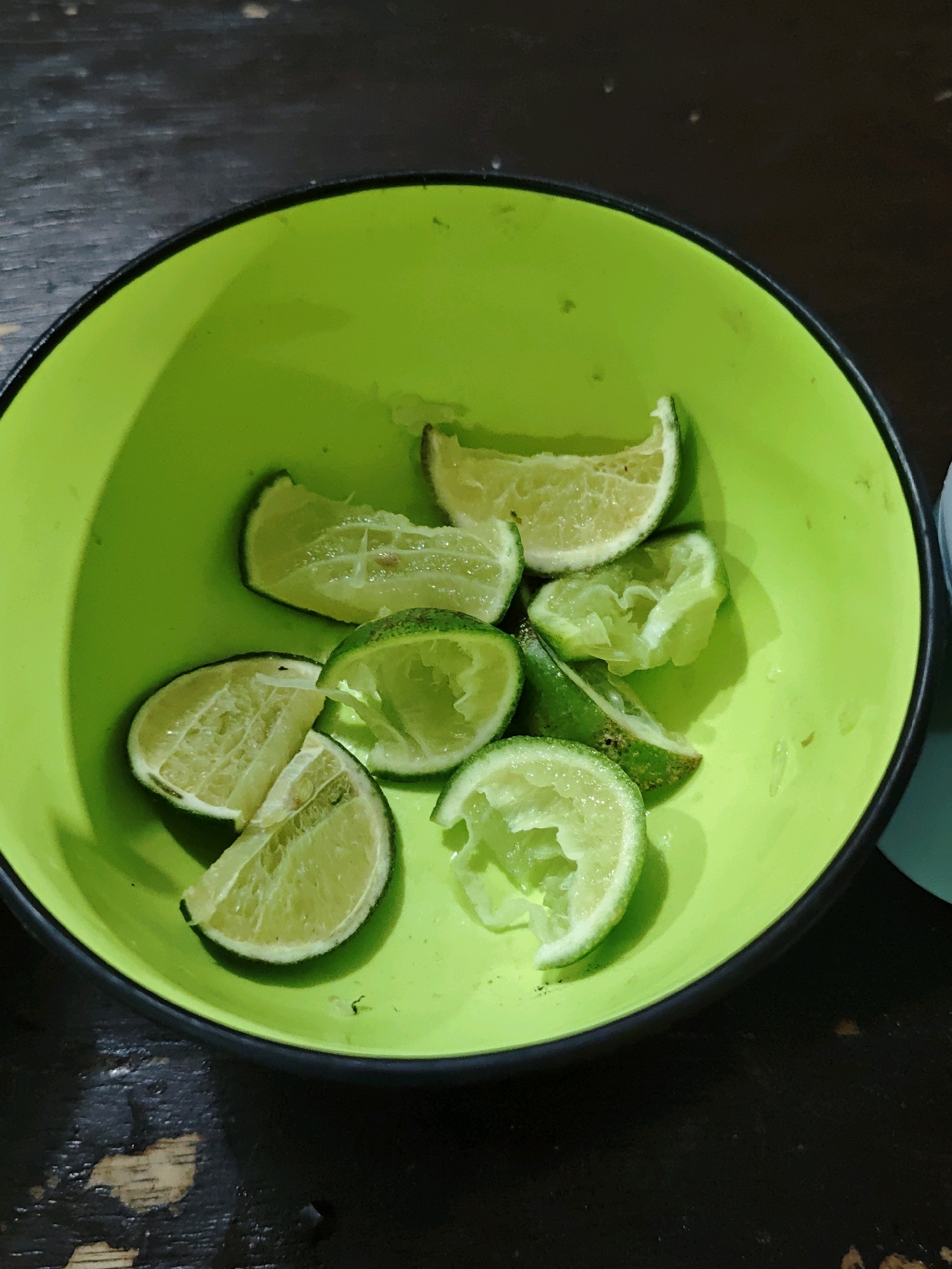 In a bright green bowl, several wedges of lime are scattered. Some wedges are whole and appear juicy, while others are partially squeezed, revealing their pulpy interiors. The bowl sits on a dark, textured surface that resembles wood, possibly a table. The lighting casts a subtle shadow and highlights the reflective surfaces of the lime wedges and the bowl. The background is dark and slightly out of focus, suggesting an indoor setting in Ngasem, Indonesia. No people are visible in the image.
