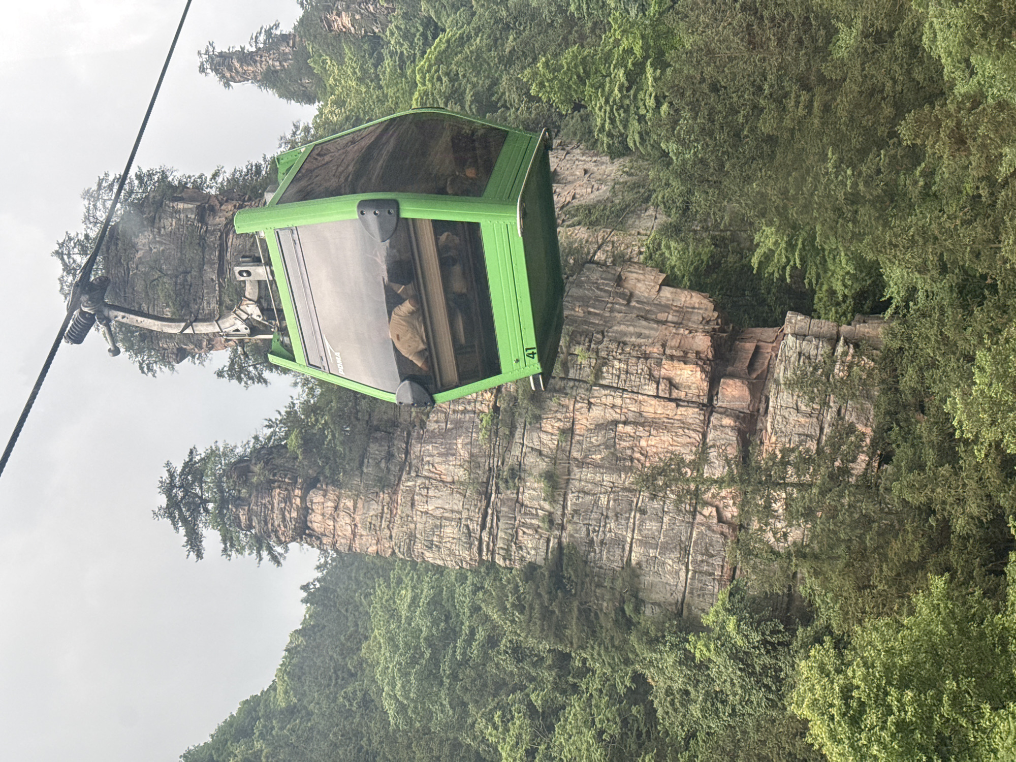 A green gondola rises above rugged rock pillars in Zhonghu, China. The gondola, filled with passengers, is suspended by a cable system. The rock formations are partially covered with greenery. The sky is overcast.